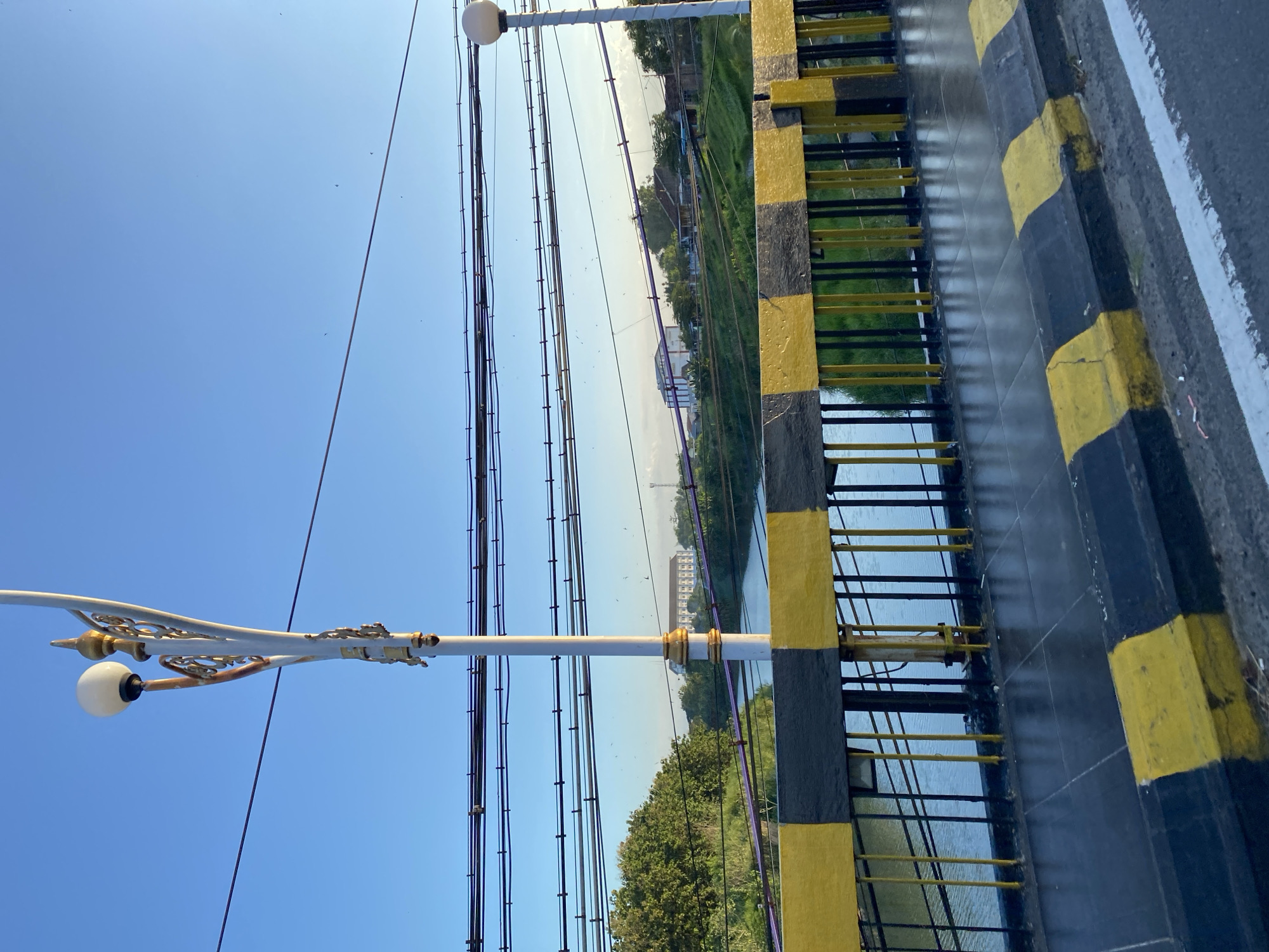 The image depicts a view from a bridge, potentially over a river or canal, in Madiun, Indonesia. A decorative lamp post stands to the left, wires and cables crisscrossing the sky. The bridge railing features yellow and black hazard stripes. A body of water is visible below, with vegetation along its banks. In the distance are buildings, possibly a school or administrative complex. The sky is clear and blue.