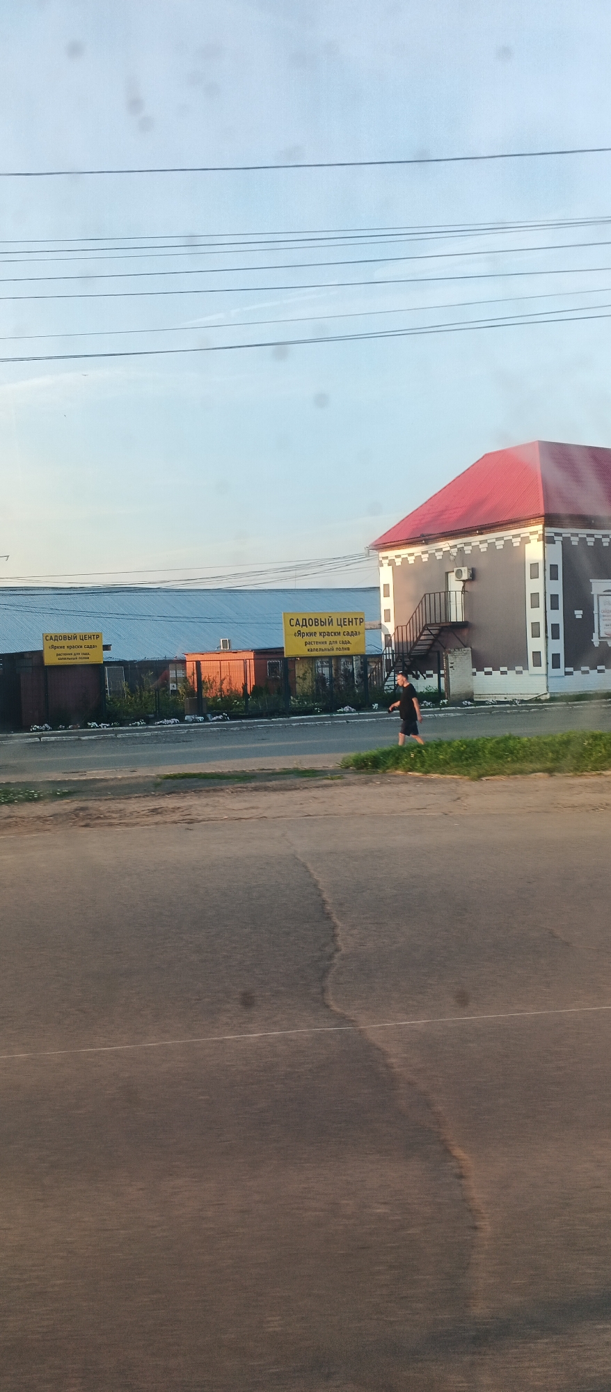 The image shows a street scene in Izhevsk, Russia. On the left, there is a long, blue-roofed building with two yellow signs that read "САДОВЫЙ ЦЕНТР" (Garden Center) and "Яркие краски сада" (Bright colors of the garden), along with "растения для сада" (plants for the garden) and "садовый инвентарь" (garden tools). To the right of this building, there is a smaller, orange-colored building. Further to the right, a light brown building with a red roof and white trim is visible. A person wearing a dark t-shirt and shorts is walking on the sidewalk in front of the brown building. The street in the foreground appears to be paved and shows some wear and tear, with white lines indicating lanes. The sky is a pale blue with visible power lines overhead. The overall scene suggests a commercial area, likely on the outskirts of the city, given the presence of a garden center.