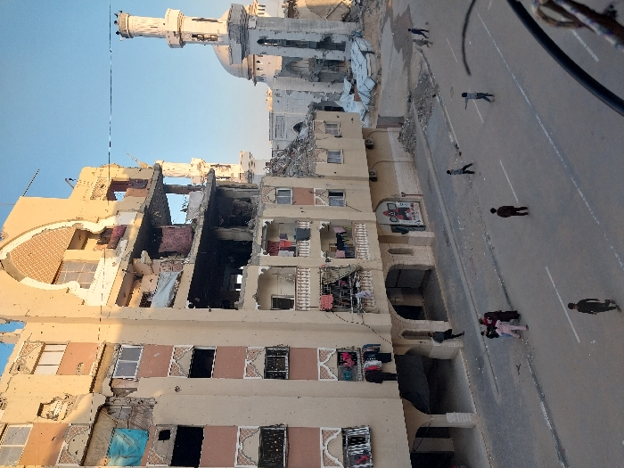 The image depicts a street in Khan Yunis, Palestinian Territory, showing a scene of extensive urban damage under a clear sky. A multi-story, light-colored residential building occupies the left side, displaying severe structural damage, including large sections of missing walls exposing interior rooms. Numerous impact marks are visible on the facade, and various items of clothing hang from balconies and exposed window openings. In the background, a mosque with a damaged minaret and dome stands amidst a wide area of rubble. The paved street below is partially covered with debris and is populated by approximately 10-12 individuals, mostly appearing as children or young adults. A small group is gathered on the sidewalk near the damaged building, while other individuals are dispersed across the street, some walking, and two on the far right appear to be playing.