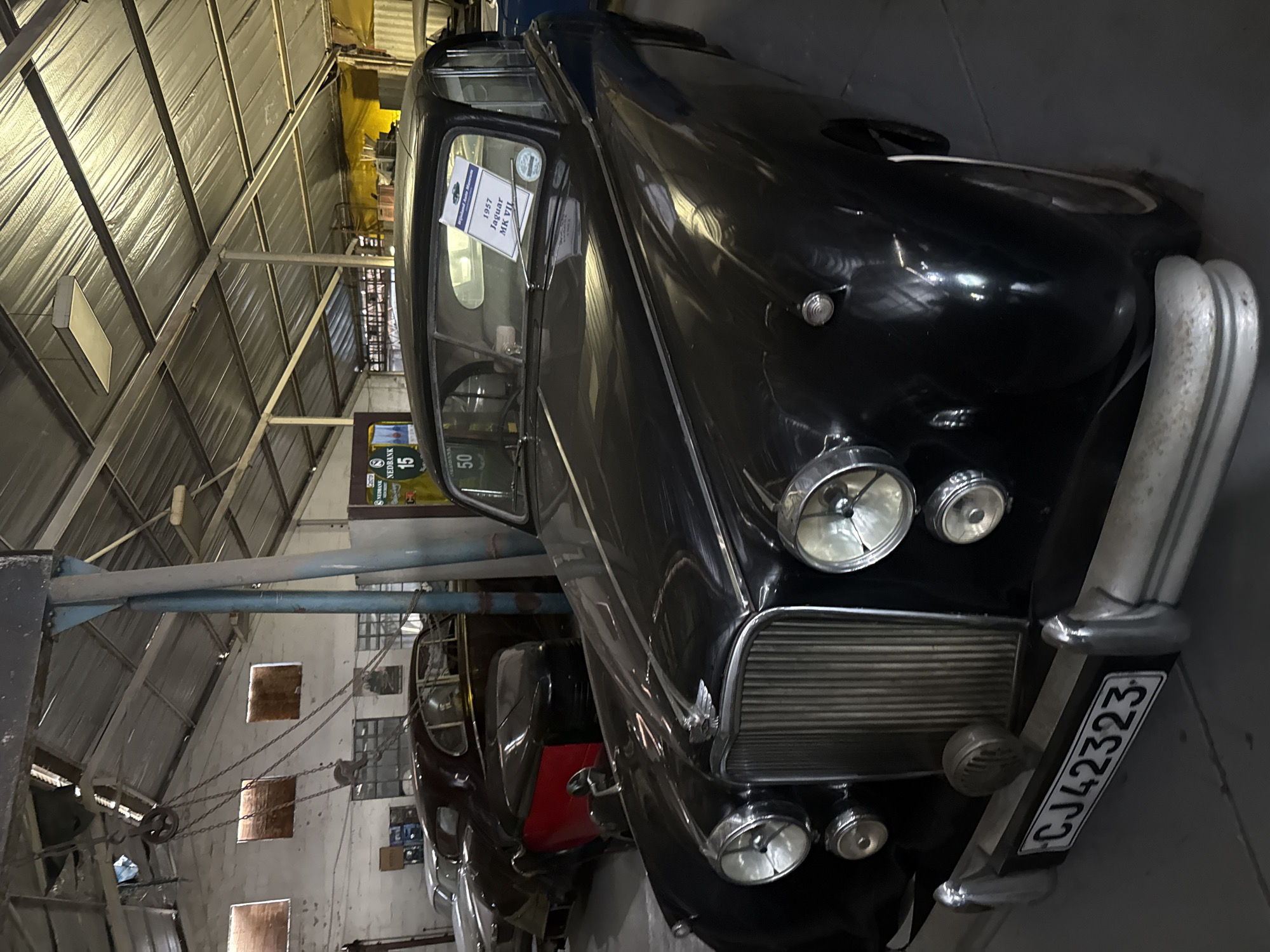 The image shows a black 1957 Jaguar MK VII car inside a building. The car has a license plate that reads "CJ42323". A sign is visible on the car's windshield, identifying it as a 1957 Jaguar MK VII. The image was taken at the Franschhoek Motor Museum in Kraaifontein, South Africa. The interior of the building shows a metal roof structure and a partially visible white wall with small, square windows. Other cars are partially visible in the background.