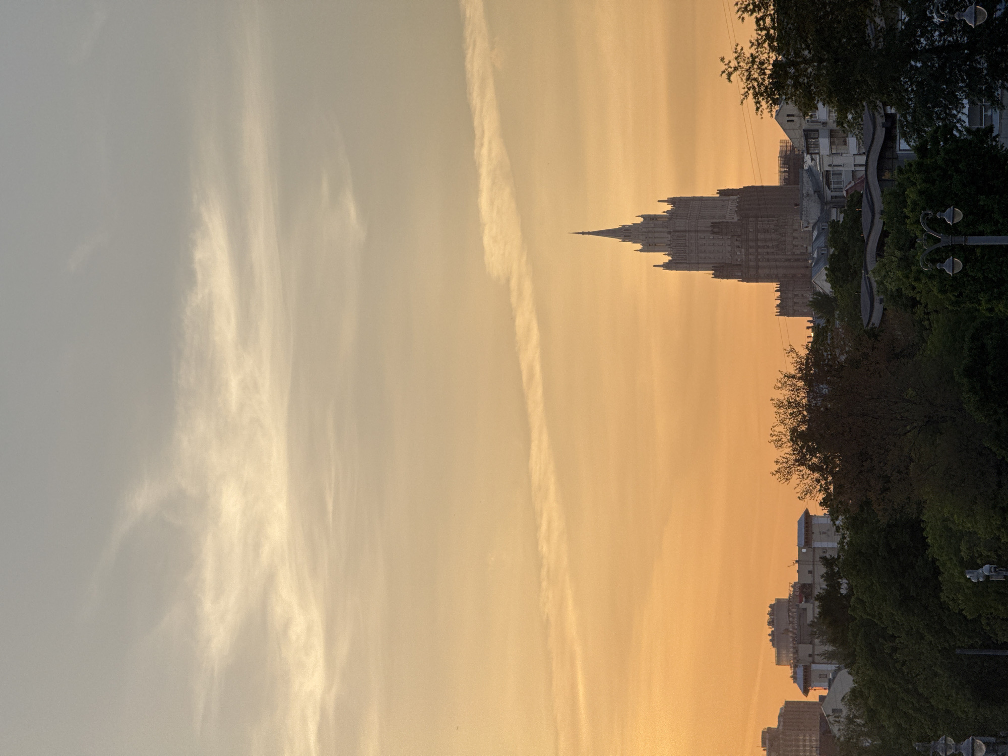The image shows the Ministry of Foreign Affairs building in Moscow, Russia, silhouetted against a yellow sky with streaks of clouds. Trees and streetlights line the right side of the frame. Other buildings are visible on the bottom right.