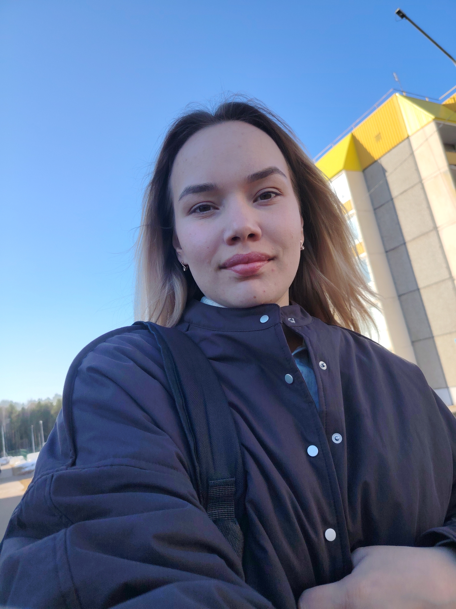 A young woman is taking a selfie outdoors on a sunny day. She has light brown hair, a light complexion, and is wearing a dark blue jacket with silver buttons. A backpack strap is visible on her left shoulder. Behind her, the sky is clear blue, and a multi-story building with yellow and light grey sections can be seen. In the distance, there are trees and possibly street lights. The scene appears to be in an urban or suburban area, though exact location Марковский is difficult to precisely ascertain based on the image alone.