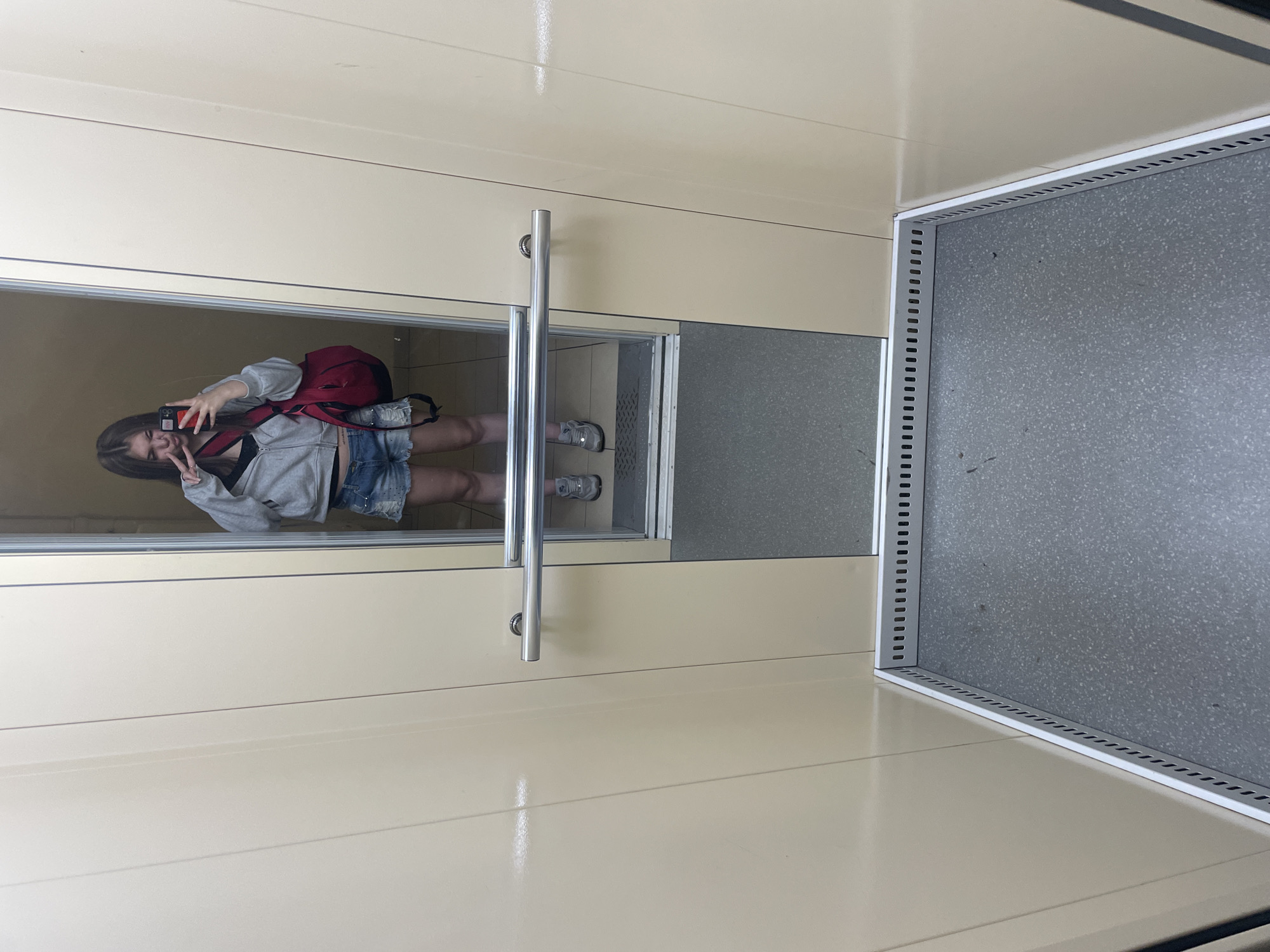 The interior of an elevator in Lednevo, Russia, features pale beige vertically paneled walls and a grey speckled floor lined with white perforated metallic trim at the base. A large, full-length mirror forms one wall, reflecting a young woman taking a self-portrait and the opposing interior. A dual silver-chrome handrail is mounted horizontally on the wall opposite the mirror.
The woman is dressed in a light grey zip-up hoodie over a black top, distressed blue denim shorts, and grey athletic shoes. She holds a red-cased smartphone in her right hand, capturing her reflection, while making a peace sign with her left hand. A red duffel bag is slung over her right shoulder. Her facial expression is characterized by slightly pursed lips.