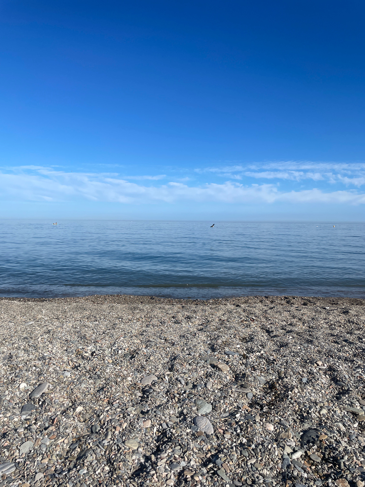 The image depicts a serene beach scene with a clear blue sky and calm water. The foreground is filled with a dense layer of various-sized pebbles and rocks, creating a textured ground. The mid-ground features the tranquil, blue expanse of the sea, with gentle ripples on its surface. In the distance, a single bird is captured in flight, its silhouette stark against the horizon. A small buoy is visible to the right, and further to the left, a boat with at least two occupants can be seen. The horizon line is distinct, separating the water from the sky, which is dotted with wispy white clouds. The overall impression is one of peacefulness and natural beauty. The location is identified as the coast near Lújar, Spain.