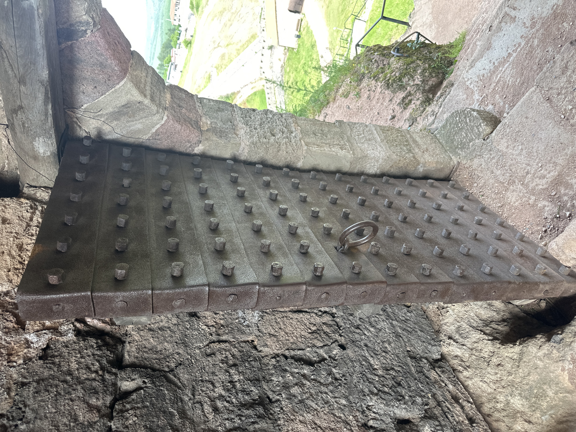 The image depicts a raised portcullis, a heavy, gated barrier, in Belogradchik, Bulgaria. The portcullis is constructed of a dark gray, possibly iron, grid of horizontal and vertical bars reinforced with numerous regularly spaced bolt heads. A metal ring is visible on its surface. The portcullis is suspended between stone walls, revealing a view of a grassy landscape and some buildings outside of the castle. The stone walls surrounding the portcullis are rough and weathered. The lighting suggests a bright day.