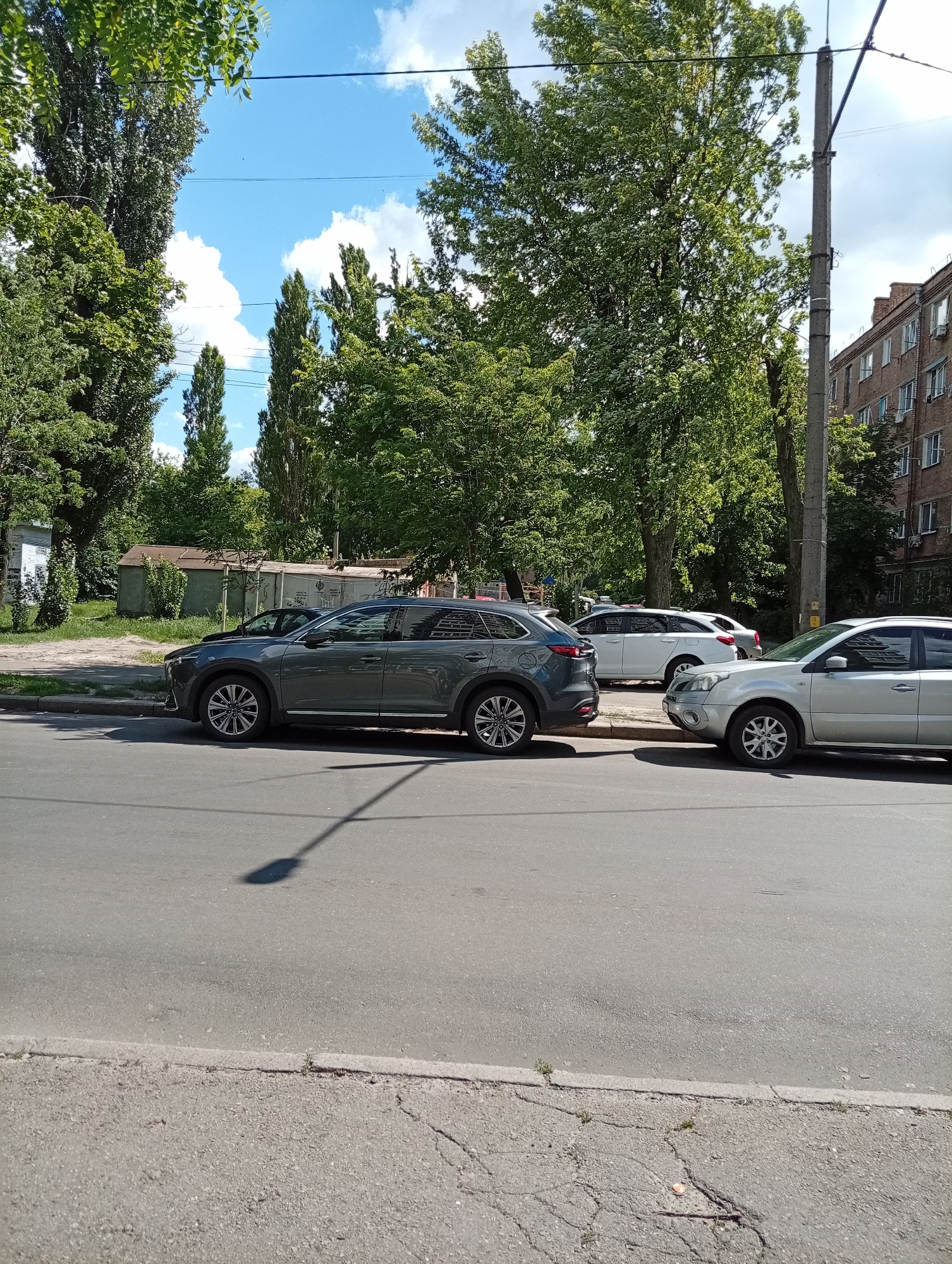 The image shows a street scene on a sunny day with a clear blue sky and some white clouds. Lush green trees line the street, and a multi-story brick building is visible in the background. Several cars are parked along the curb. In the foreground, a gray SUV is parked. Next to it, a silver sedan is parked, and further down the street, a white SUV is visible. The scene appears to be a residential or urban area, possibly a street in a city or town. The time of day seems to be daytime, given the bright sunlight casting shadows. There is no visible text in the image. The overall impression is a calm, everyday scene.