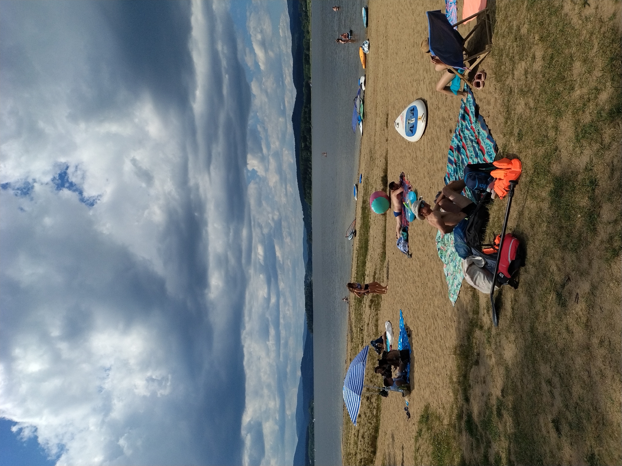 The image depicts a sunny day at a beach with a large body of water and hills in the background, suggesting a location like the Radslav Reservoir in the Czech Republic. Several people are present on the sandy beach, some of whom are sunbathing on towels and blankets. An umbrella provides shade for a small group of people. Various water sports equipment, including paddleboards and a large inflatable ball, are scattered on the sand. The sky is filled with dramatic clouds, indicating a dynamic weather system. The overall atmosphere is one of leisure and outdoor recreation.