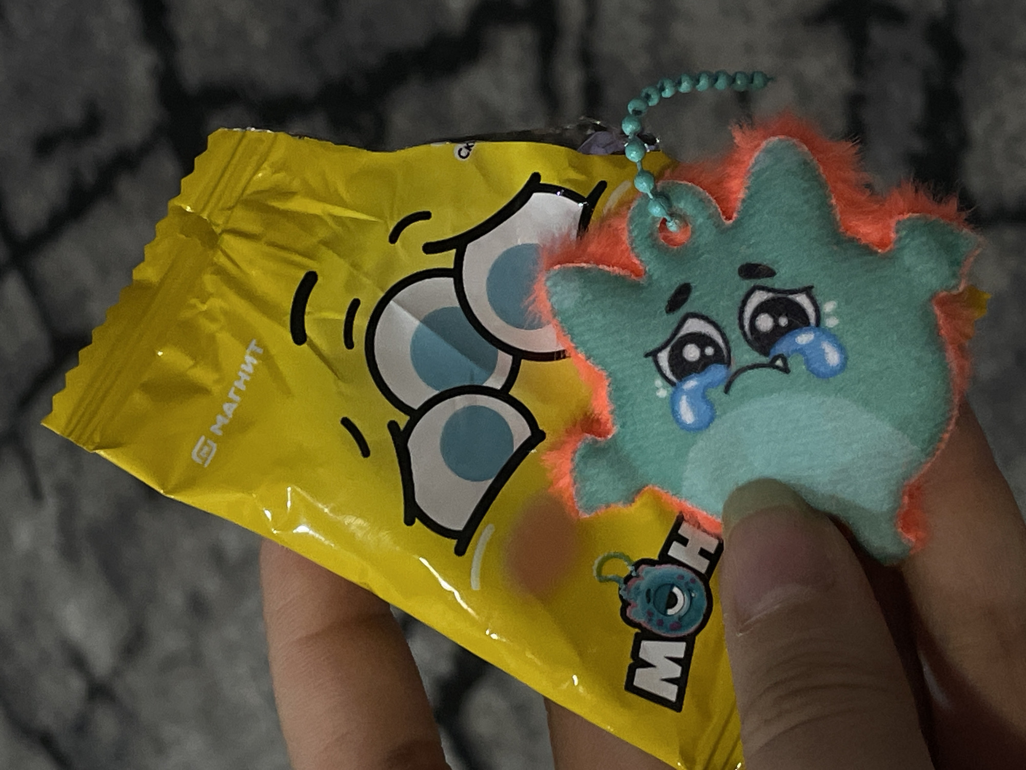 A close-up shows a hand holding a yellow package bearing cartoon-like eyes and the word "MOHOW" in black and white lettering, along with the word "Магнит" in Russian. Attached to the package is a small, fuzzy, turquoise plush toy with an orange trim, depicting a crying character with large eyes and teardrops. The image was taken in the city of Chaikovsky, Russia.
