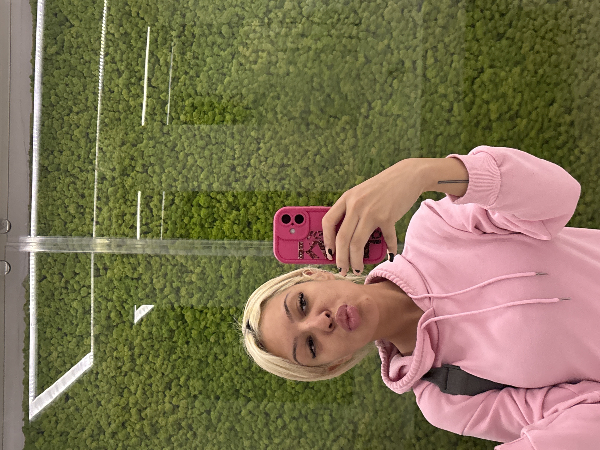A woman stands in front of a moss-covered wall, holding a pink cell phone in her right hand, while taking a selfie. The woman has short blonde hair and is wearing a pink hoodie. The moss wall is contained within a clear frame or enclosure and has white accents in a few spots. The city and specific location are unknown.