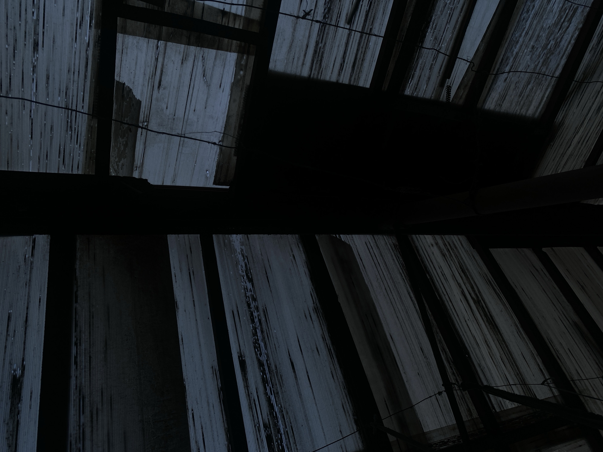 The image depicts an abstract view of a partially collapsed wooden structure, possibly a roof or ceiling, with visible beams and planks. The scene is dimly lit, with deep shadows creating a sense of decay and disrepair. The texture of the weathered wood is prominent, showing horizontal lines and variations in color from dark gray to lighter, almost white patches. Thin wires or strings are visible, hanging loosely in some areas. The overall impression is one of abandonment and neglect. There are no people or specific identifiable objects within the frame. The location in the city of Brest, Belarus, is not discernible from the visual information provided.