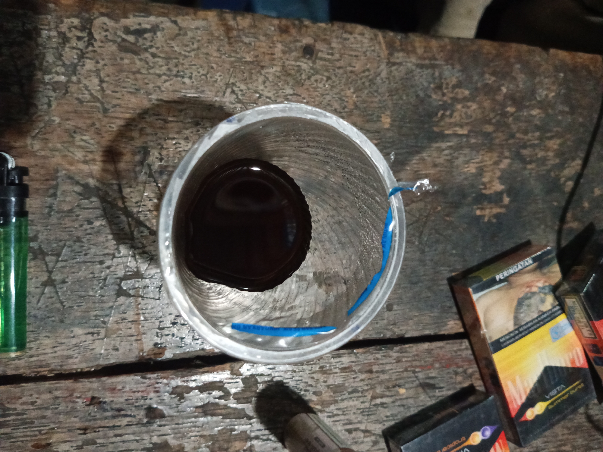 The image depicts a close-up, overhead view of a weathered wooden surface, likely a table, in Tumampua, Indonesia. A clear plastic cup containing a dark liquid, possibly coffee or tea, is centrally positioned. The interior of the cup shows some residue, and blue tape is wrapped around the exterior. To the left of the cup, a green disposable lighter is visible. To the right, several packs of cigarettes are arranged, including one with a prominent orange and yellow design and the word "VISTA" and "Summer Burst" visible, alongside another pack with a warning label in Indonesian. The overall scene suggests a casual, perhaps outdoor or rustic setting.