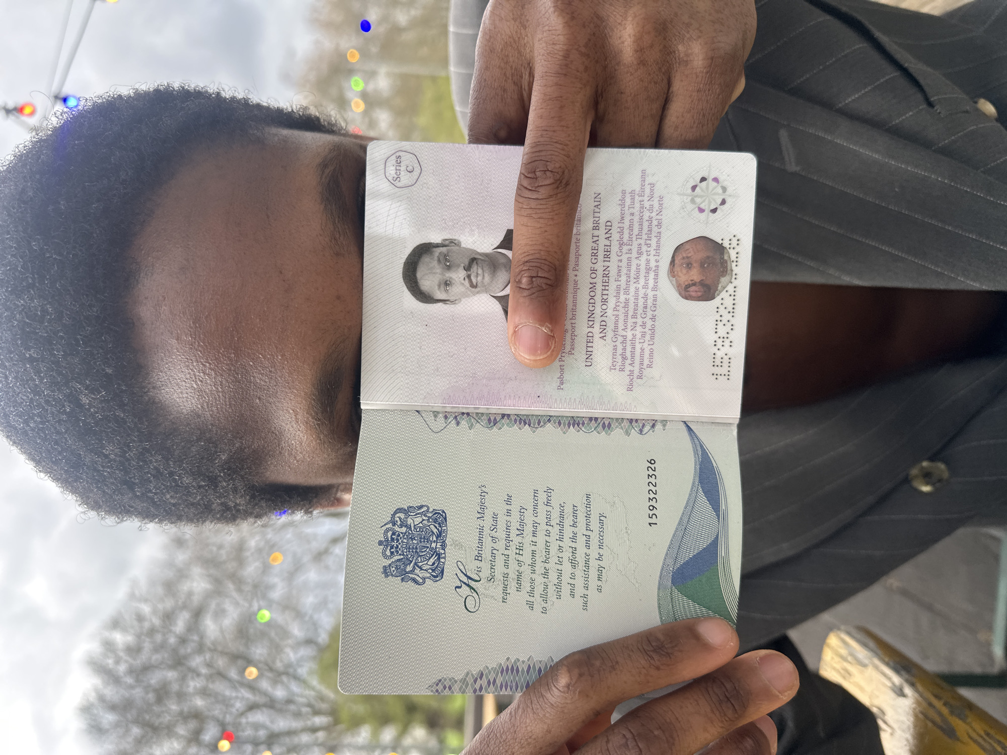 A medium-shot captures a person holding open a United Kingdom passport, presumably on display for verification or identification. The person's face is partially visible, with their forehead and hairline in clear view, showcasing a closely cropped haircut. Their skin tone is medium to dark brown. The person is wearing a grey pinstripe suit jacket. The passport is held open to display the photo page and the facing page with official text. The text includes the phrase "United Kingdom of Great Britain and Northern Ireland" in English and other languages. A serial number is visible on the right page. In the background, blurred shapes and colors suggest an outdoor setting, with what appears to be string lights or decorations. The context suggests the photo was taken in a public location in the United Kingdom.