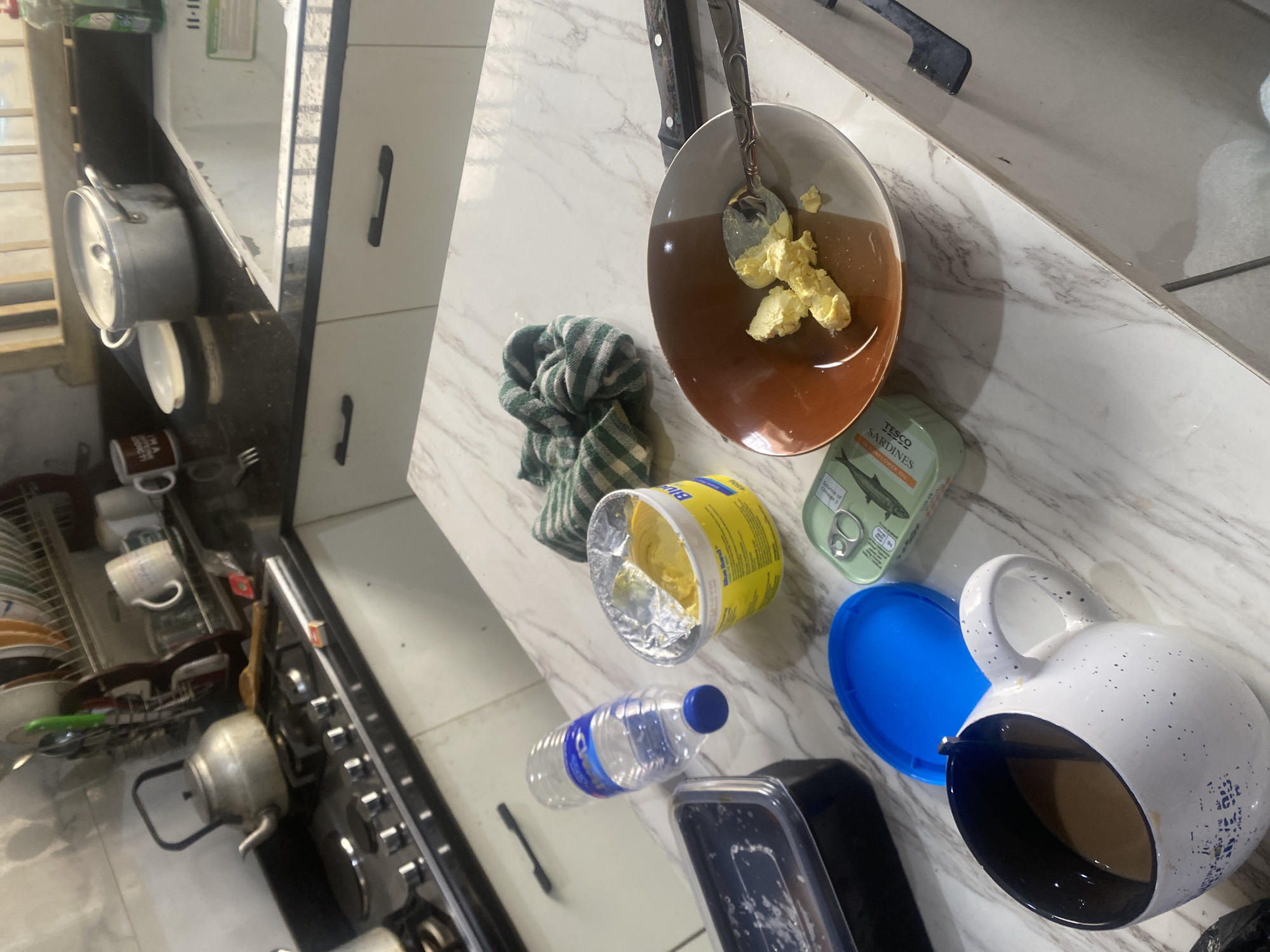 The image depicts a countertop scene in a kitchen located in Lagos, Nigeria. The countertop is a light marble. Visible items include a bowl with brown liquid and butter, a can of Tesco sardines in sunflower oil, a container of Blue Band margarine, a blue plastic lid, a spotted white mug with dark interior, a partially empty water bottle, a black and silver food container, a green and white striped cloth, utensils, knives, pots, dishes, cups, a kettle, and a stove. The location is identifiable by the presence of the Tesco sardines and Blue Band margarine, commonly available in Lagos, Nigeria. No people are directly visible.