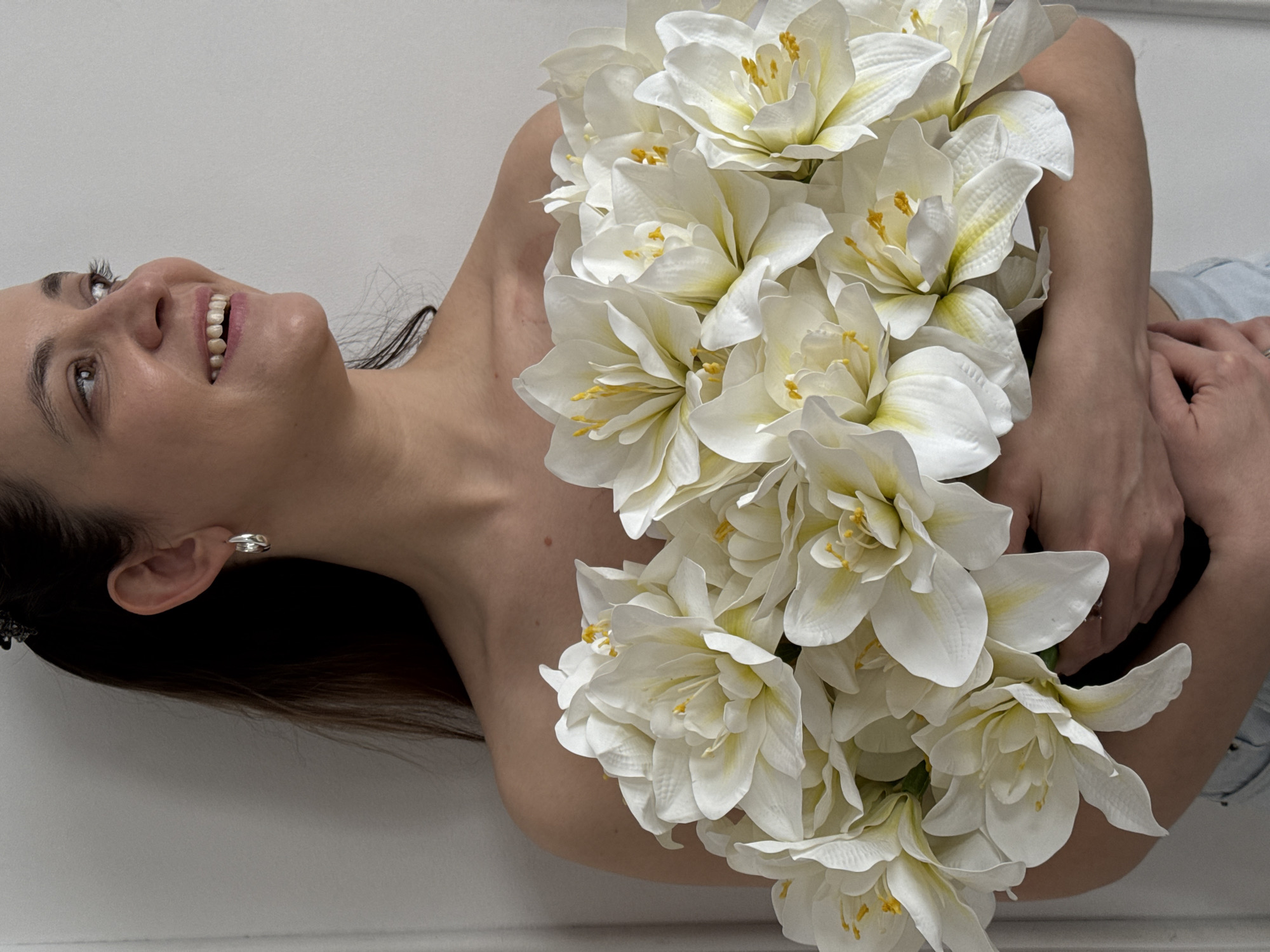 A young woman is depicted in a portrait, positioned as if reclining or lying down, with her head tilted back and looking upwards. She has a joyful expression, smiling broadly, with her mouth slightly open revealing her teeth. A large, lush bouquet of white flowers, characterized by multiple petals and yellow stamens, is held against her chest and shoulders, covering much of her upper body. Her bare skin is visible around her neck, shoulders, and arms as she cradles the flowers. She has dark hair, and a small silver-colored hoop earring is visible on her left ear. Her left hand, adorned with a ring, is seen holding the flowers near her upper arm. The background is a plain, light-colored wall, providing a simple and uncluttered setting. The overall mood conveyed is one of happiness and serenity. The photograph is taken indoors, likely in Chaikovsky, Russia, under soft, even lighting. There is no visible text in the image.