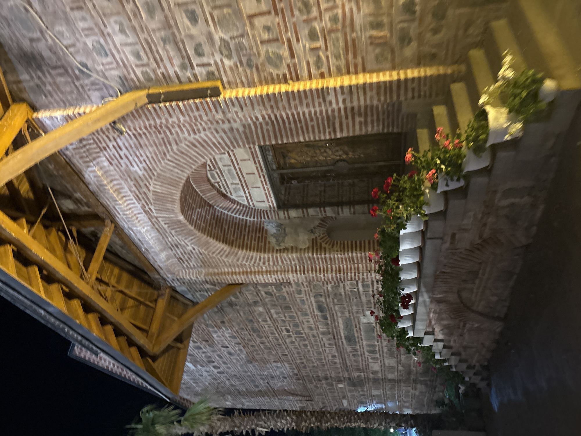 This is an eye-level, rotated photograph of the ceiling and surrounding area of the Hisar Kapia (Eastern Gate) in Plovdiv, Bulgaria. The ceiling and wall are primarily brick, with varying geometric patterns, including stripes, squares, rectangles, and an arch. The ceiling features a square opening, and a wooden support structure intersects the scene. On the right side, there is a staircase decorated with white flower pots containing flowering plants. The lower portion of the image reveals a tree and what appears to be a structure. The photograph was taken at night or in a dimly lit area.