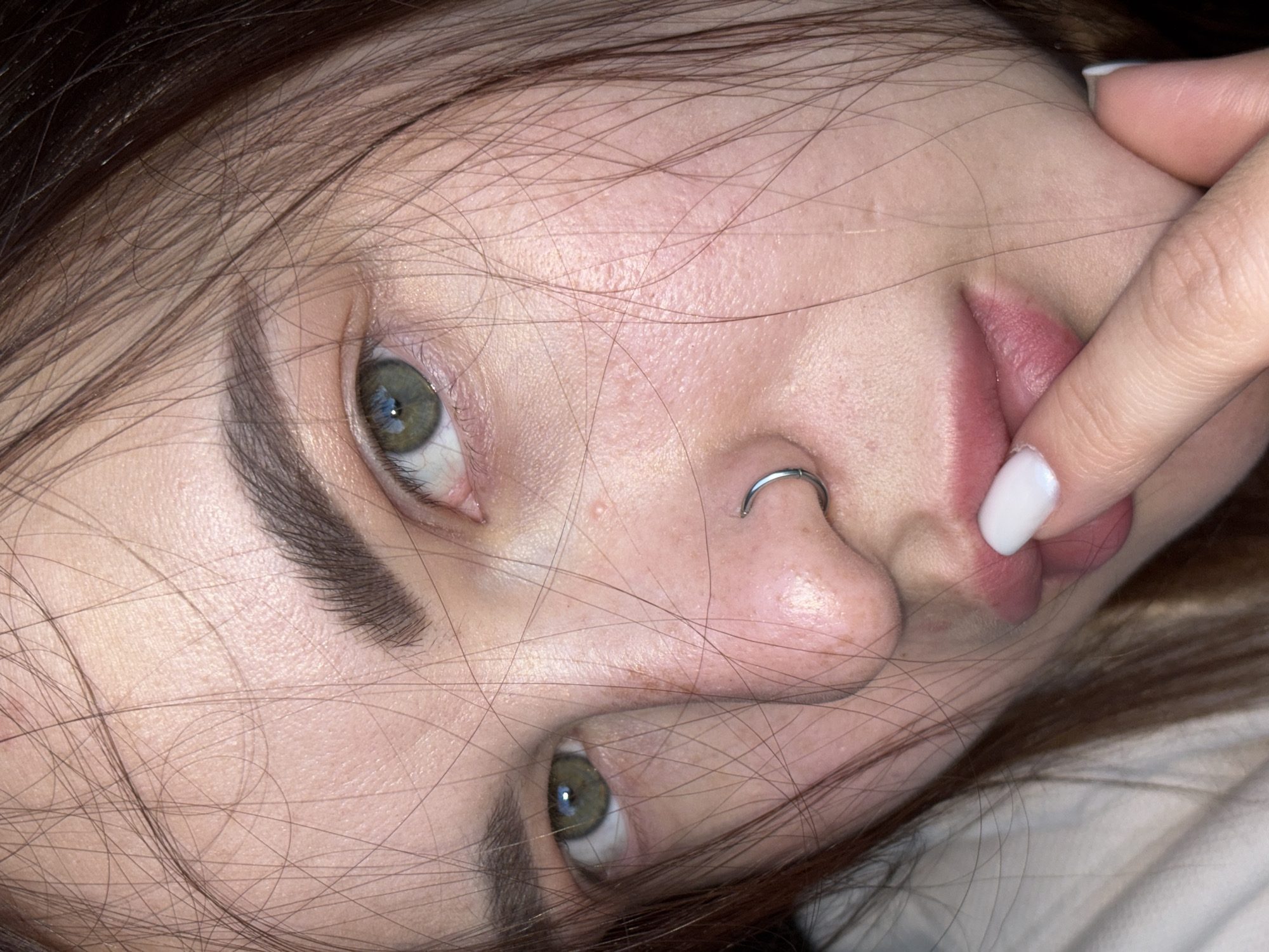 A close-up, tilted, and angled photograph depicts a woman's face. Her skin is fair, and her brown hair frames her face, obscuring portions of it. She has a nose ring on her left nostril. Her eyes are green. Her right index finger is touching her lips. The location is unspecified; no indication of Krasnodar, Russia, is present in the image.