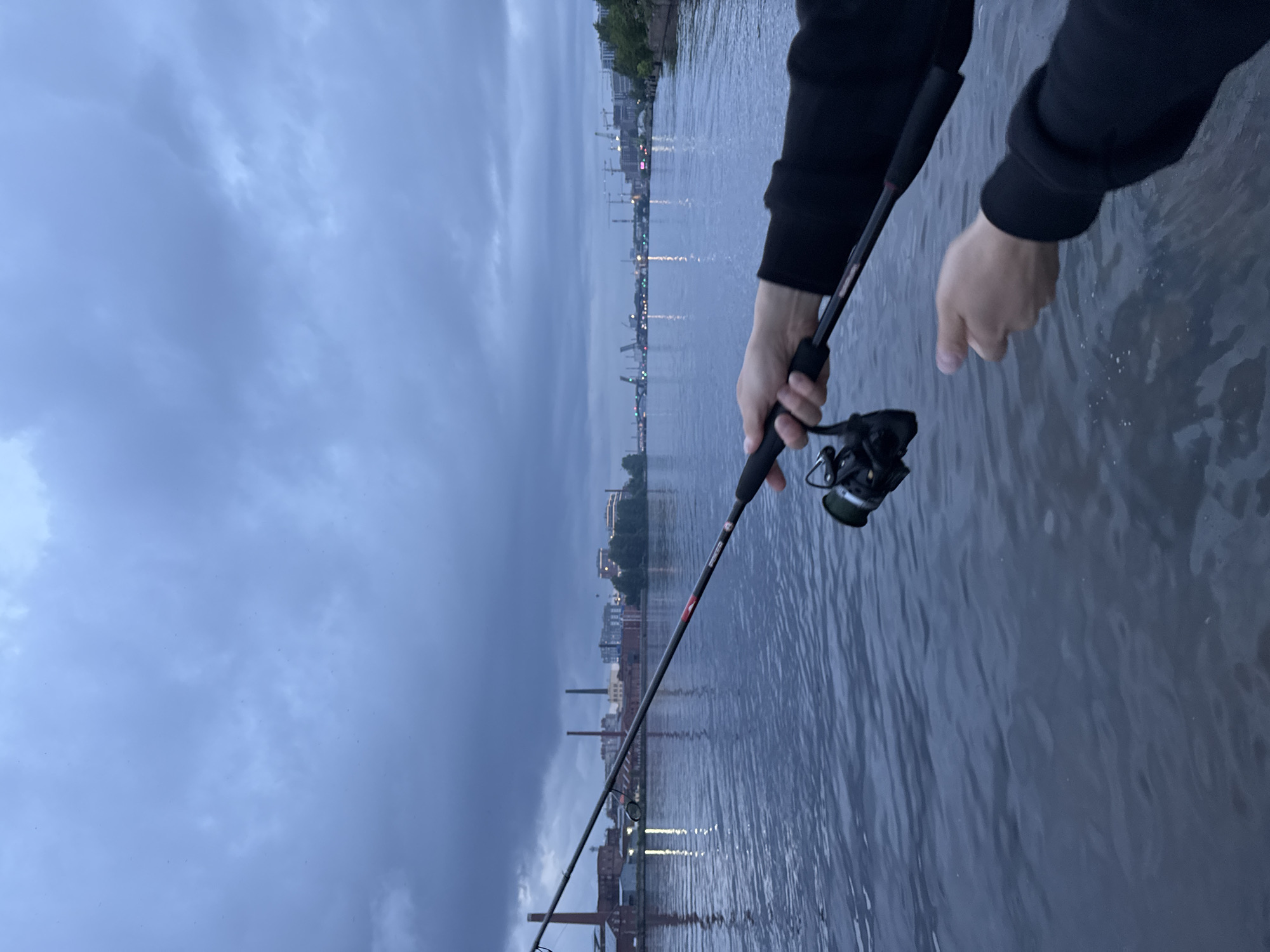 A person's hands are visible, holding a fishing rod over a body of water. The fishing rod is dark with red accents and a fishing reel is attached. The water is dark and rippling. In the background, there is a cityscape with buildings and industrial structures, including tall chimneys. The sky is overcast with dark clouds. The scene appears to be a river or canal within a city, likely St. Petersburg, Russia, given the presence of industrial architecture and potential bridge structures in the distance. The overall atmosphere is somber and dusky, suggesting either early morning or late evening.