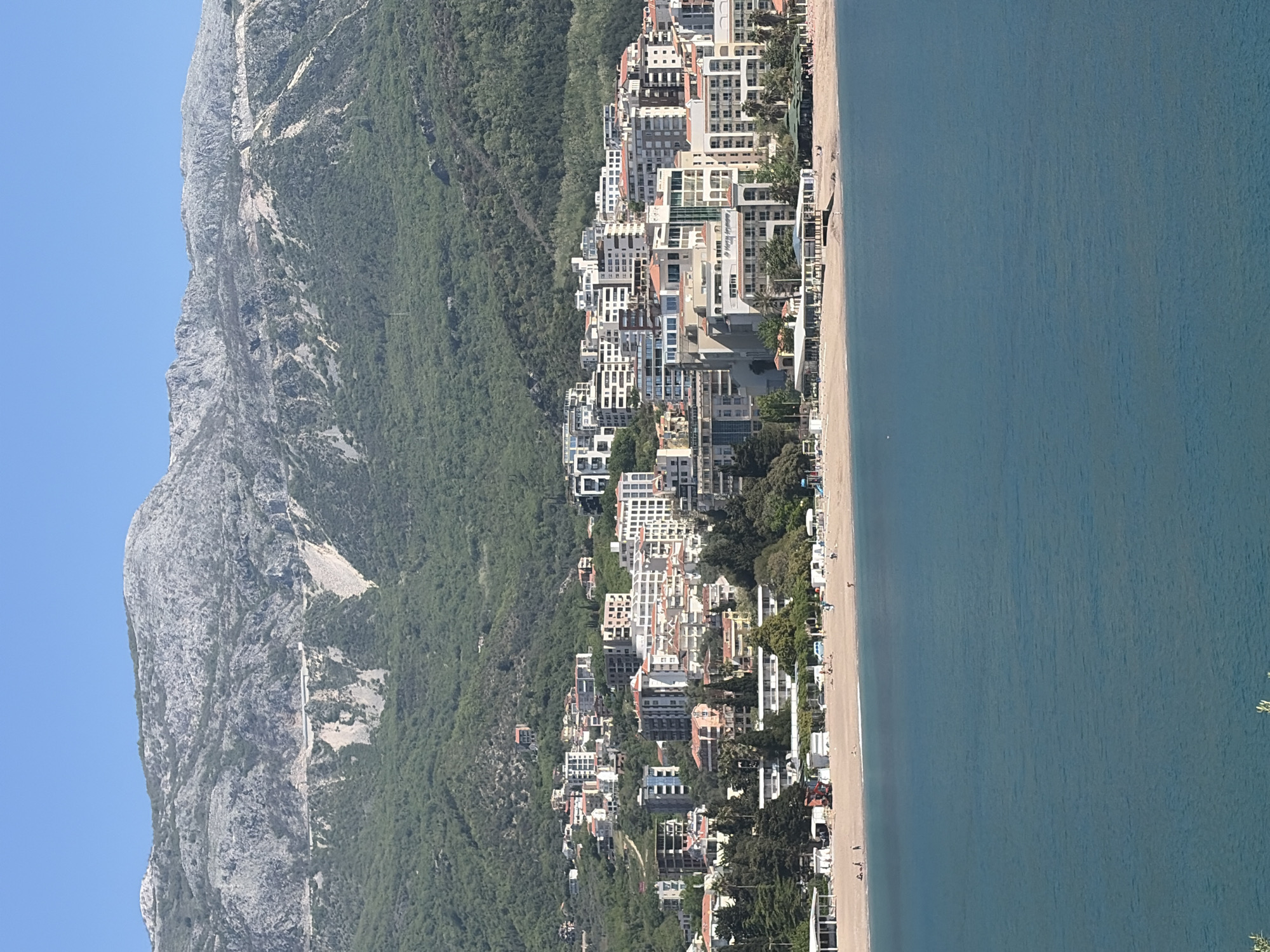 The image depicts a coastal scene in Budva, Montenegro. A large, rocky mountain covered with green vegetation dominates the left side. A dense collection of buildings and houses lines the coastline, transitioning from the mountain towards the beach. The beach is sandy with visible figures of people. The right side is filled with the blue sea. The sky is clear and blue.