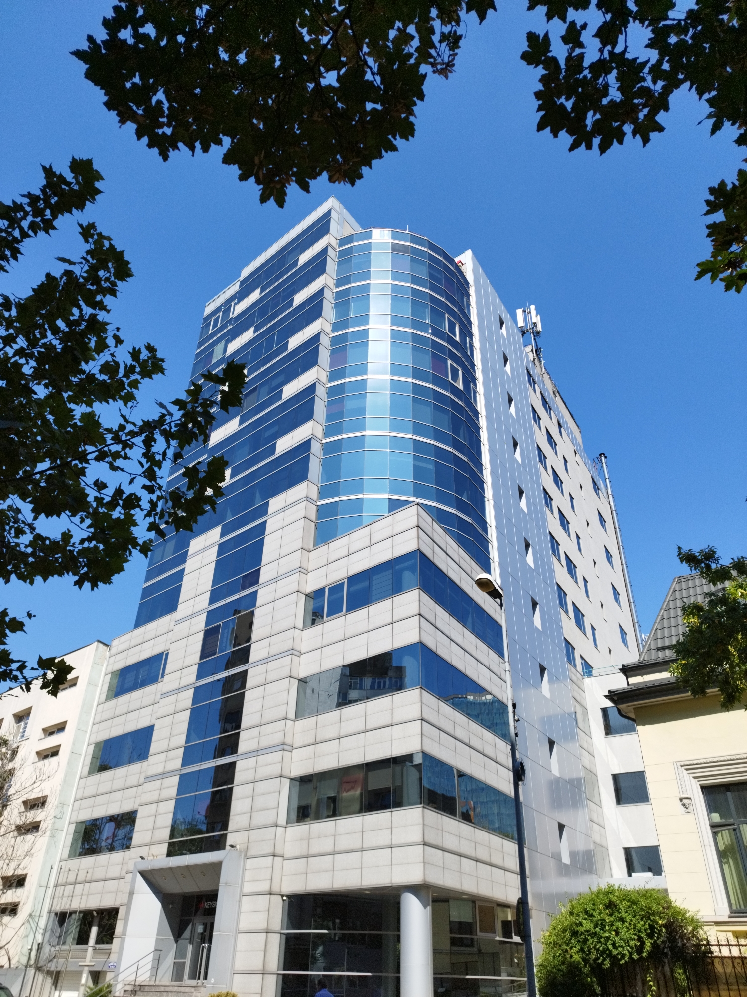 The image shows a multi-story modern office building located in Bucharest, Romania, with a bright blue sky as the backdrop. The building features a prominent curved section with a facade of blue glass windows, interspersed with white panels. The rest of the building's exterior is primarily white with rectangular blue glass windows. Several tree branches with green leaves frame the top and sides of the image, obscuring portions of the sky and building. A street lamp is visible in the foreground, to the left of the building. A smaller, older yellow building with a gray tiled roof is partially visible to the right of the main office building. Green bushes are also present in the foreground, on the right side of the image. The overall scene depicts an urban environment with a contrast between contemporary architecture and natural elements.
