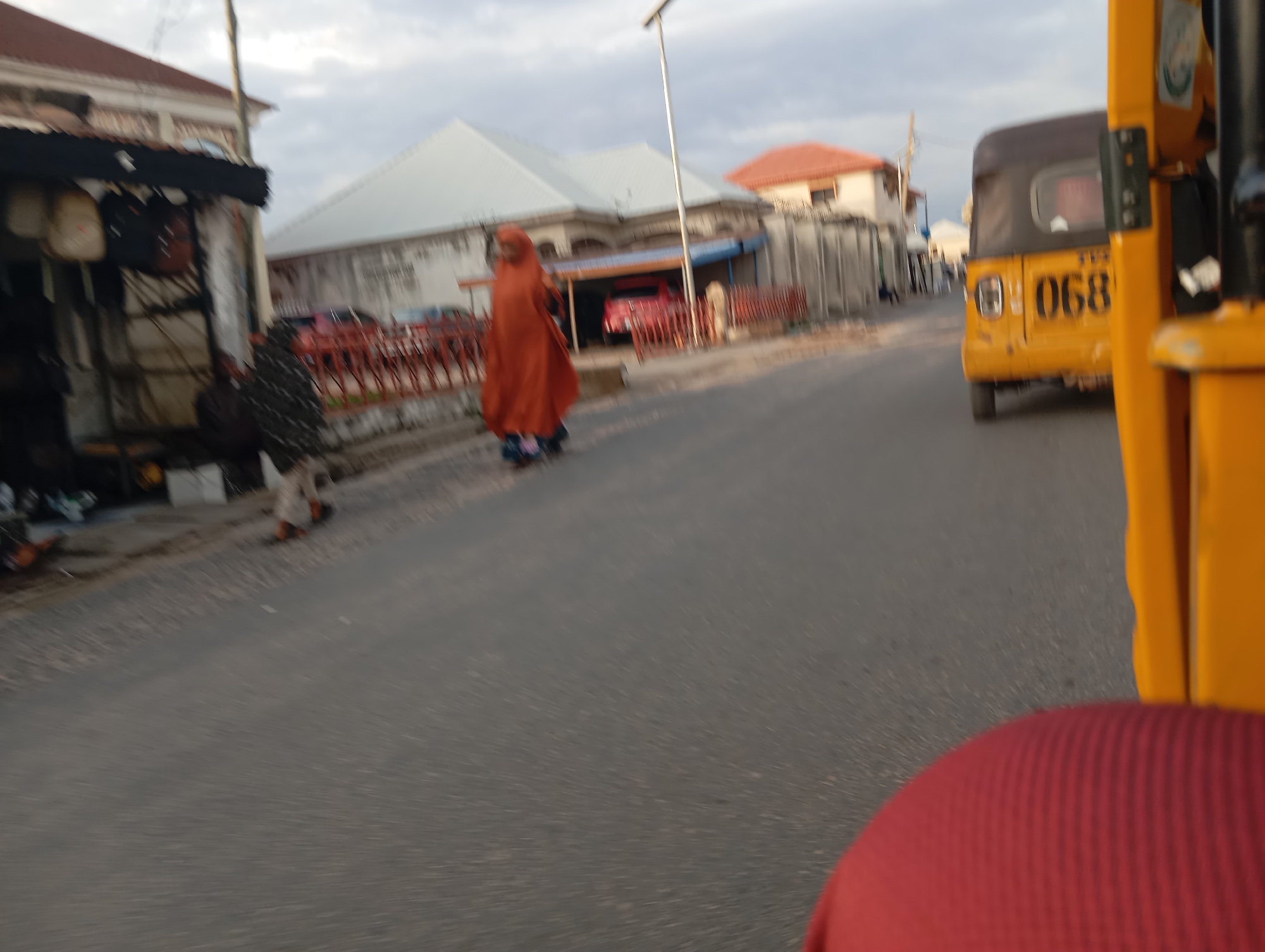 The image shows a street scene in Jalingo, Nigeria, with a blurred view of buildings, a person in an orange robe, and a yellow three-wheeled vehicle. The person, walking on the right side of the road, is partially obscured by their clothing, and their face is not visible. The yellow vehicle, a common mode of transport in Nigeria, is seen with the number "068" clearly displayed on its rear. The background reveals typical urban architecture, with buildings featuring light-colored walls and corrugated iron roofs. The presence of a street lamp and a paved road indicates an urban environment. The overall impression is a snapshot of daily life in a Nigerian city, captured from a moving perspective.