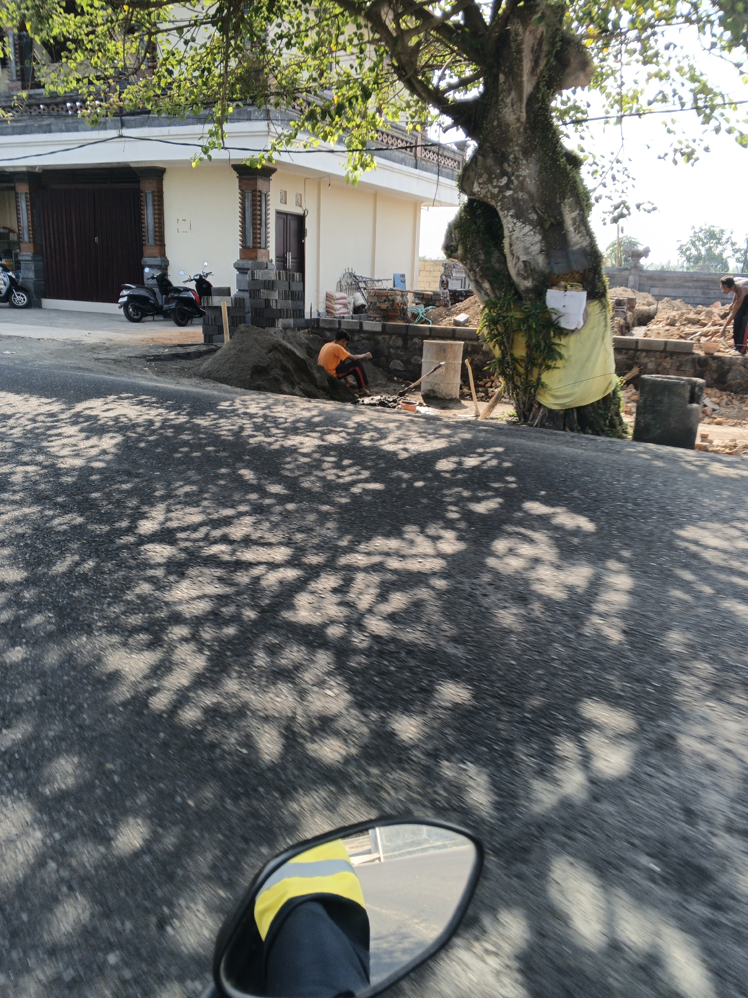 In Abiansemal, Indonesia, on a sunny day, a man in an orange shirt is working on a construction site near a large tree. He is partially obscured by a pile of sand and appears to be shoveling or moving materials. Behind him, construction work is underway, with piles of dirt, bricks, and building materials visible. A large tree with moss and vines growing on it stands prominently, draped with a yellow cloth. In the background, a building with a closed garage door and a second story balcony is visible, with two scooters parked in front. The foreground shows a road with a motorcycle's mirror reflecting the scene, including a person wearing a yellow and grey jacket. The road is dappled with sunlight filtering through the leaves of an unseen tree.