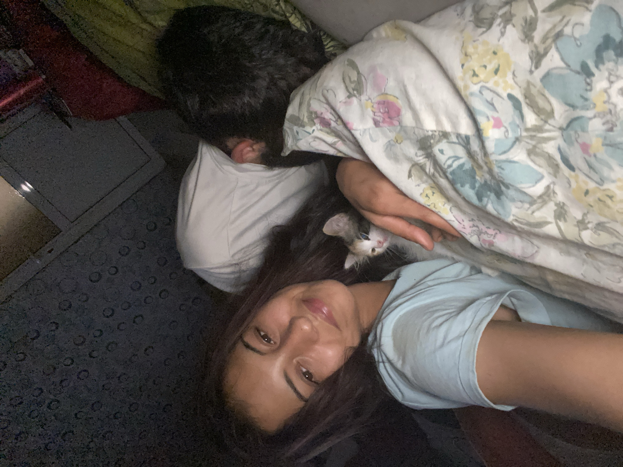 This indoor image captures a cozy scene featuring two people and a kitten. In the foreground, a woman with long, dark hair and a light blue t-shirt is looking directly at the camera with a gentle smile. She is positioned diagonally in the frame, suggesting a selfie.
Behind her, partially obscured, is another person, likely male, with dark hair, resting or sleeping with their head turned away, largely hidden by a patterned duvet. This person is wearing a white t-shirt.
Between the two individuals, a small, white kitten with black and grey patches on its face is nestled, looking towards the camera. The kitten appears to be resting comfortably.
The subjects are on what appears to be a bed, covered by a light-colored duvet featuring a floral pattern with pale yellow, light blue, and pink elements. A white pillow is visible under the head of the second person. The lighting is dim, indicating it is likely evening or night. To the left, a dark, textured surface, possibly a carpet or mat, and a dark rectangular object are visible. The overall mood of the image is peaceful and affectionate. There is no visible text in the image.