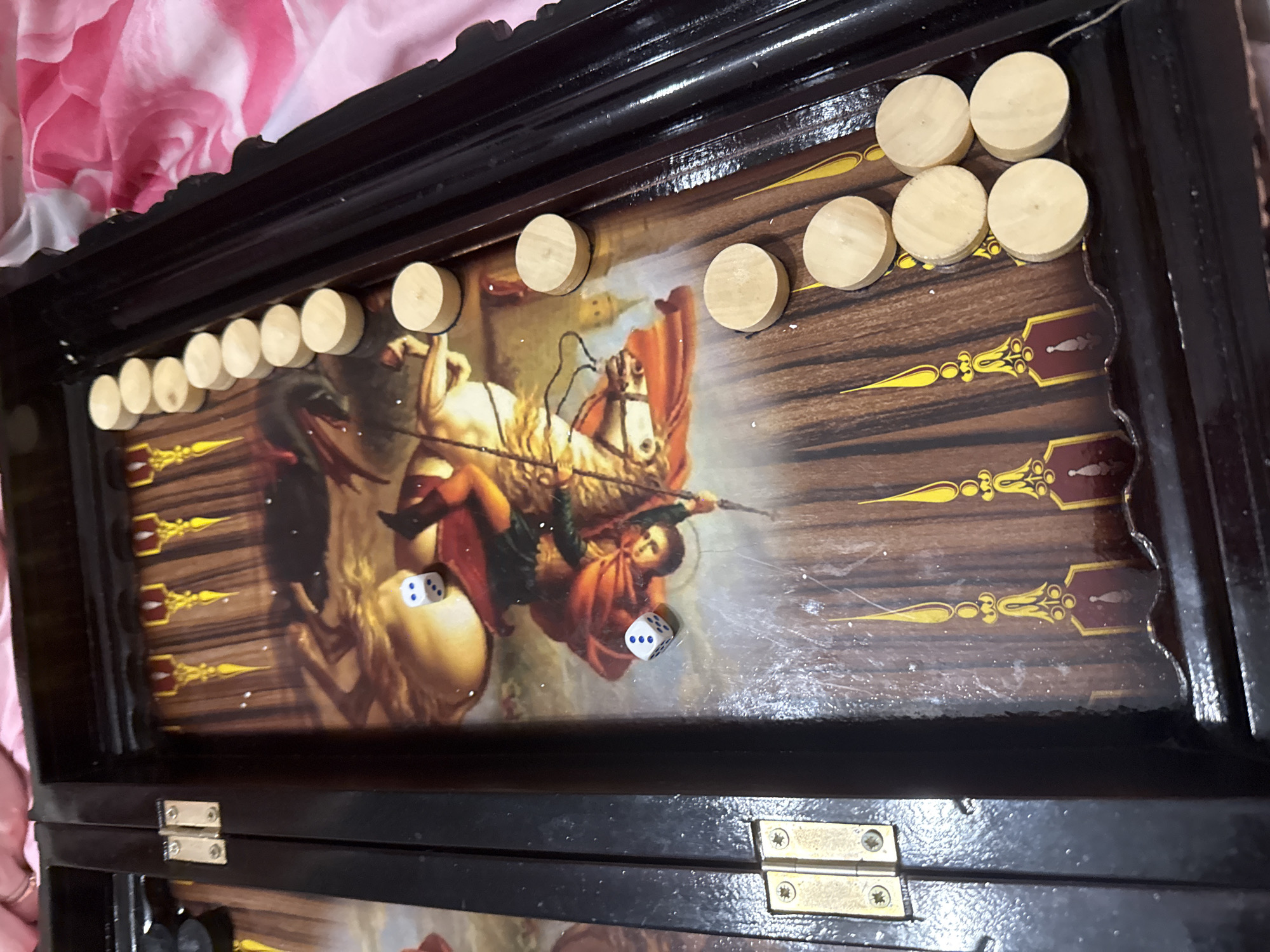 An open backgammon board is depicted from an overhead angle, set for play. The board itself is dark, possibly black or dark brown, with a glossy finish. Its playing surface features a printed image of a classical painting, centrally showcasing a figure on horseback, wielding a spear, engaged in a dramatic scene that suggests a conflict or slaying. The figure is adorned in a prominent red cloak, and the horse is light-colored and rearing.
Numerous light-colored, circular wooden checkers are arranged along the top edge of the board and positioned on some of the dark points (triangular playing areas) which are decorated with gold and dark red patterns. Two white dice with blue pips are visible on the playing field; one shows a face of one pip, and the other shows three pips. The board's dark, framed edge reveals brass hinges towards the bottom. The entire arrangement rests on a soft, patterned fabric featuring pink and white floral designs. The setting is indoors, with the activity of a backgammon game either in progress or freshly set up. The provided context indicates the location as Khimki, Russia, though no specific visual cues within the image confirm this. No visible text is present.