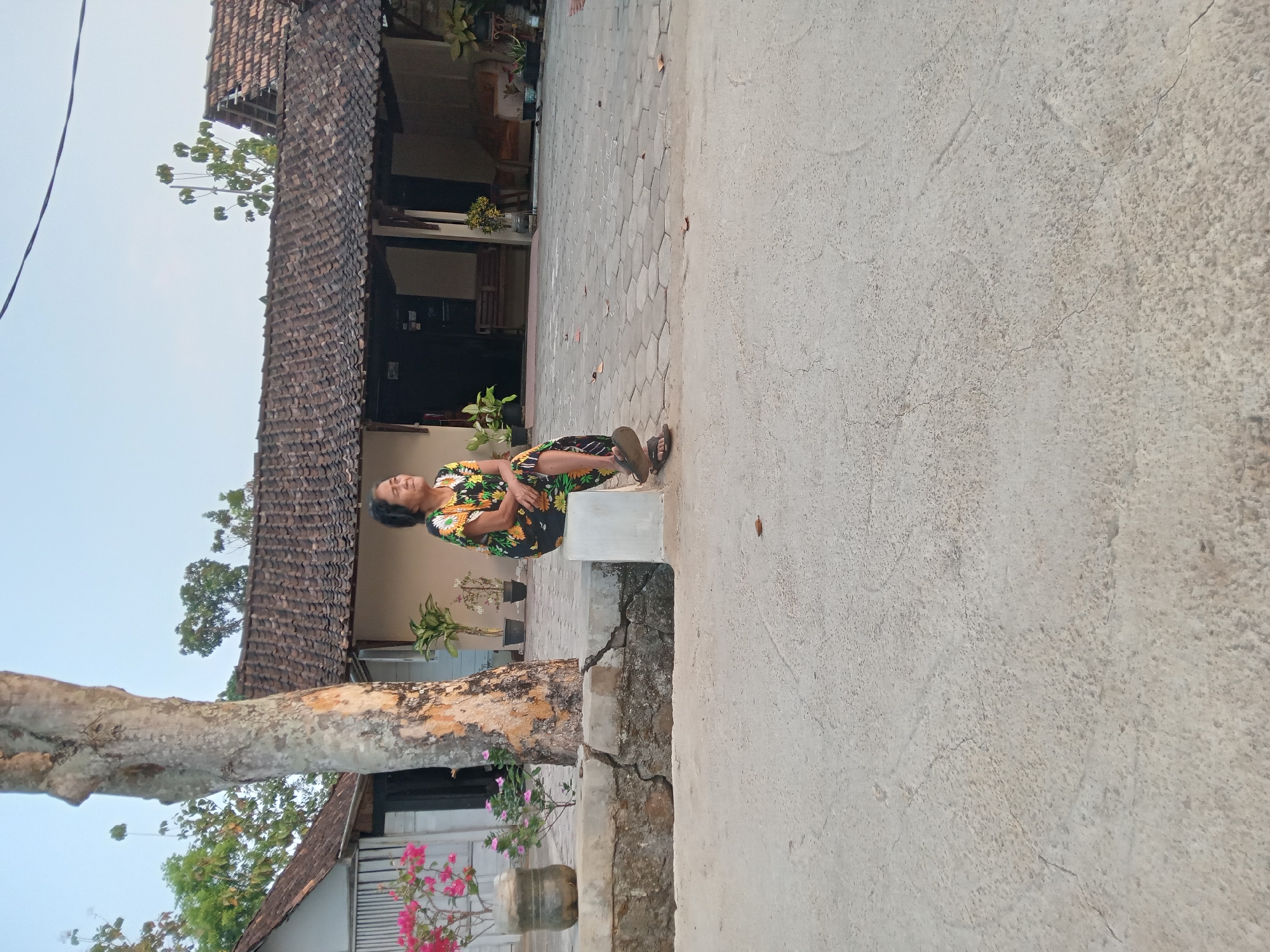 A medium shot captures a woman sitting on a concrete block in front of a single-story building with a tiled roof in Kedunggudel, Indonesia. The woman, who appears to be of Southeast Asian descent, is wearing a brightly colored floral dress and flip-flops. She is looking to her right with her eyes closed. A large tree trunk stands to her left, with a cluster of pink flowers in a recycled plastic bottle planter to the left of the tree. In the background, the building features a covered patio area with wooden benches. The ground in front of the building is paved with interlocking hexagonal stones, and a concrete path extends to the foreground. The sky is clear and light blue, suggesting it is either early morning or late afternoon. The overall impression is one of a peaceful, everyday scene in a rural Indonesian setting.