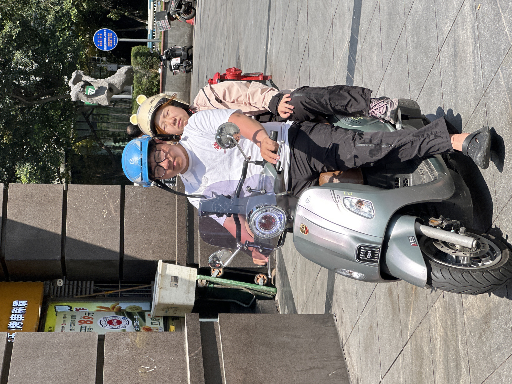 Here is a detailed description of the image:
**General Description:**
The image shows two people riding a silver SUNRA scooter on a paved area next to a building. The scene appears to be outdoors during daylight, with clear and bright lighting.
**People:**
1. **Rider (presumably male):** Wearing a light blue helmet, glasses, and a white t-shirt. He is sitting on the scooter and seems to be operating it.
2. **Passenger (presumably female):** Wearing a helmet with bear-ear decorations, a light pink jacket, and black pants. She is reclining on the rider, appearing relaxed or possibly asleep.
**Objects:**
1. **Scooter:** A silver SUNRA scooter is prominently featured. Details like the front wheel, headlight, mirrors, and license plate are visible.
2. **Helmets:** Both individuals are wearing helmets.
3. **Building:** A building is partially visible on the left side of the frame. Its architecture includes stone blocks.
4. **Signage:** Chinese text signage is visible. A blue sign reads "Please consciously park in the marked area" and "The direction of the car head should be unified and super positive."
5. **Other Scooters:** Other scooters are parked behind the featured scooter.
6. **Advertisement:** An advertisement for food is visible. The text on the sign reads "Alipay group purchase authority, the wheel shines, kebab hegemony, 8.9 yuan, Alipay sweep."
**Actions/Interactions:**
1. **Riding:** The scooter is either stationary or in the process of starting to move, with the rider seemingly in control.
2. **Reclining:** The passenger is reclining on the rider, suggesting closeness or rest.
**Environment:**
1. **Location:** The scene seems to be in an urban area with paved surfaces and adjacent buildings.
2. **Lighting:** The scene is brightly lit, suggesting a sunny day.
3. **Vegetation:** Trees are visible in the background.
**Location in Yuzhong District, China:**
Without precise geolocation clues within the image, it is difficult to pinpoint the exact location within Yuzhong District. However, based on the architecture, scooter, style, and signage, it is likely a public space, such as a sidewalk or designated scooter parking area, in a commercial or residential zone of Yuzhong District, China.