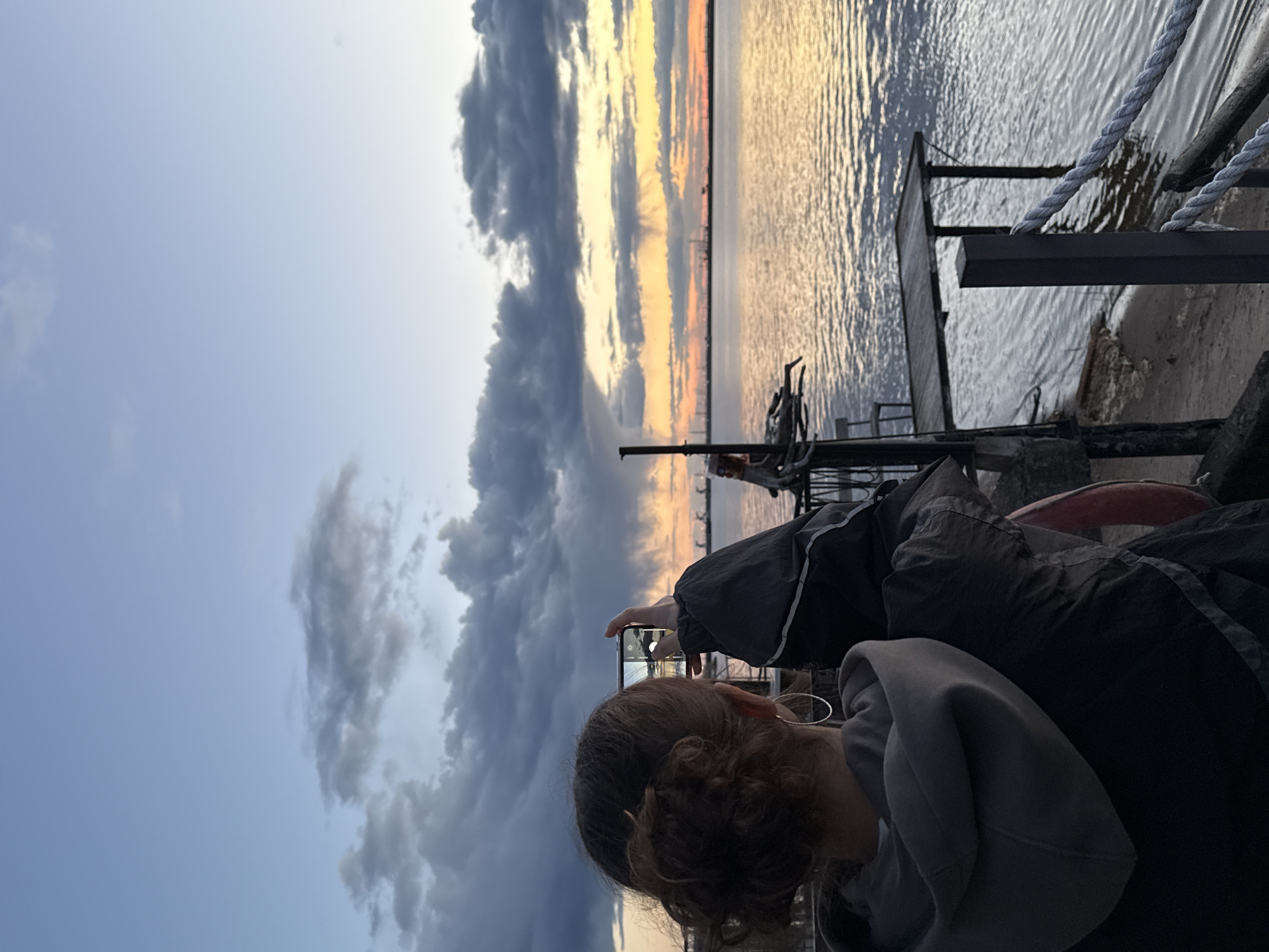 A woman with curly brown hair is holding a smartphone up to take a picture of a sunset over a body of water. She is wearing a gray hoodie and a black jacket. The sky is cloudy, with dark clouds on the left side and orange and yellow hues on the horizon. There is a dock or pier with railings and ropes in the background. The location is Chaikovsky, Russia.