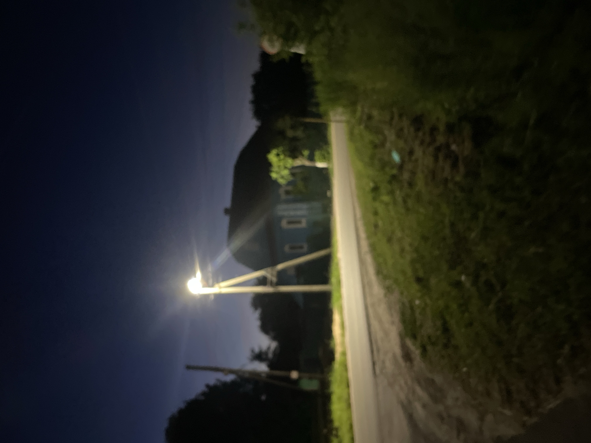 The image depicts a dimly lit rural scene at night in the village of Krasnyye Baki, Russia. A street lamp illuminates a paved road and a grassy embankment. In the background, a blue wooden house with visible windows stands adjacent to dark trees. The overall atmosphere is quiet and rural, with no people or vehicles visible.
