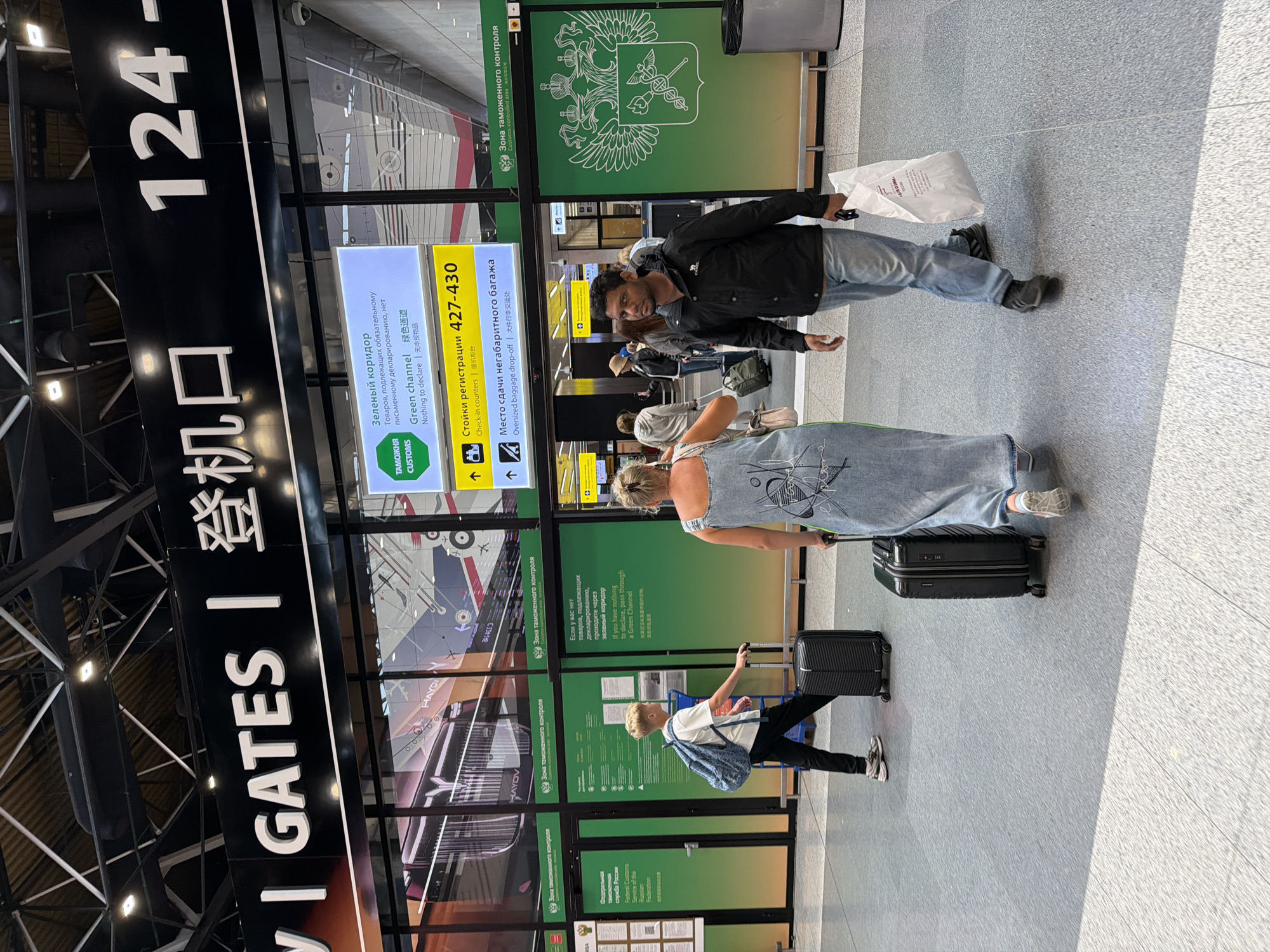 An interior shot captures a bustling airport scene in Khimki, Russia, likely Sheremetyevo International Airport, identified by the signage in Russian and English, as well as the Russian coat of arms displayed prominently. The image focuses on two individuals walking across the concourse. In the foreground, a man wearing a black jacket and blue jeans is walking towards the viewer's left, carrying a white paper bag in his right hand. To his left, a woman in a denim dress is also walking towards the left, pulling a black suitcase behind her. Behind them, other travelers and luggage are visible, indicating a busy travel environment. Overhead, large signs display gate information and directional arrows in multiple languages, including Russian, Chinese, and English. Green signage indicating customs areas, specifically a "Green channel" for those with nothing to declare, is also visible. The floor is a speckled grey tile, and the ceiling features exposed industrial beams and lighting fixtures. The overall atmosphere is one of movement and transit, typical of an international airport.