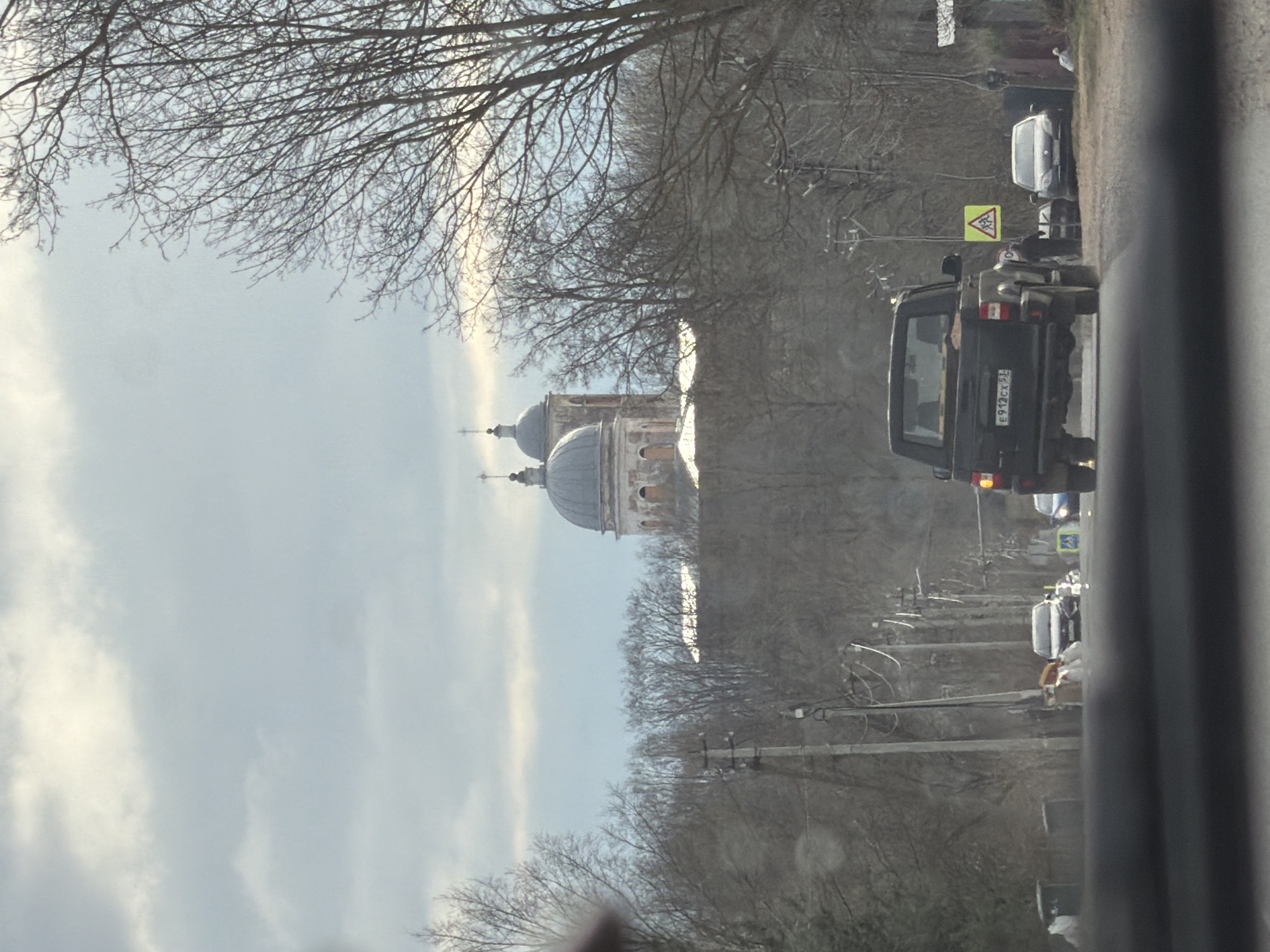 The image, taken in Бронницы, Russia, shows a partial view from inside a vehicle, likely a car. A road is visible, with several vehicles traveling on it. A black SUV with license plate "E972CX 50" is immediately in front of the photographer's vehicle. Further ahead, other cars are visible, along with a pedestrian on the roadside. On the left side of the road, a large building with distinctive domes is present. Bare trees line the road. A "Children" sign is visible on the right side of the road. The sky is overcast. Utility poles are also visible alongside the road.