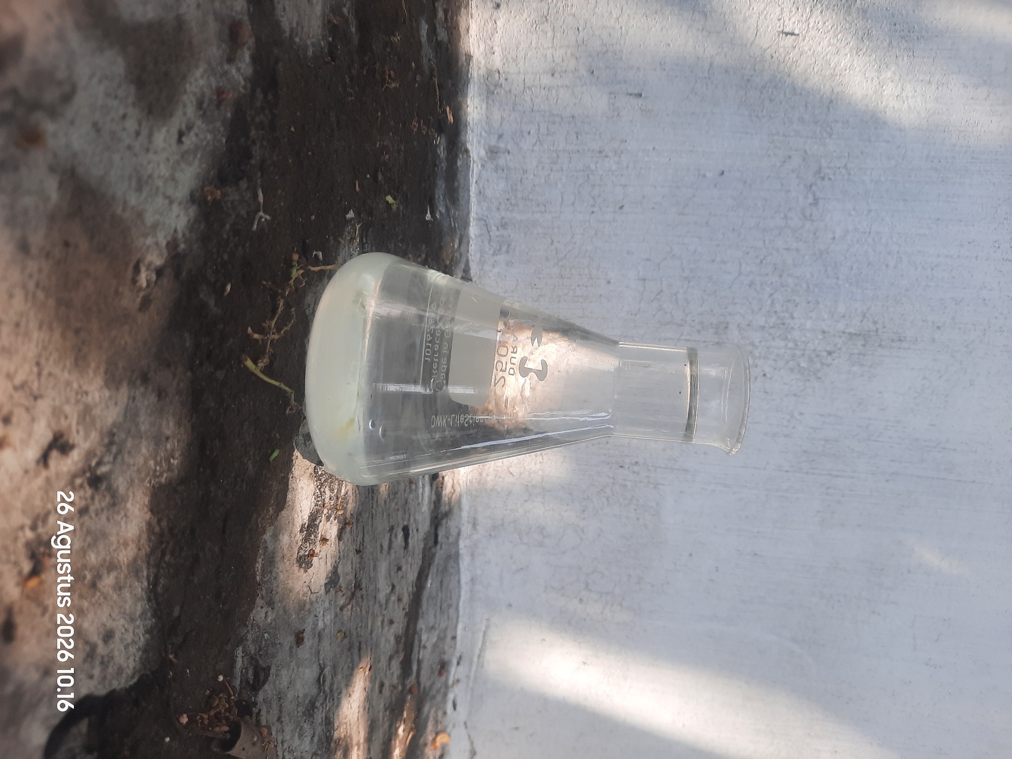 An Erlenmeyer flask containing clear liquid with a milky-white precipitate at the bottom sits on a coarse, dark-colored surface. The flask is illuminated by sunlight, casting shadows from nearby foliage onto a textured, off-white wall in the background. The surface beneath the flask appears to be a mixture of soil and debris, with dry leaves and small twigs scattered around. In the bottom right corner, a timestamp reads "26 Agustus 2026 10.16", indicating this photograph was taken in Gending, Indonesia. No individuals are present in the scene.