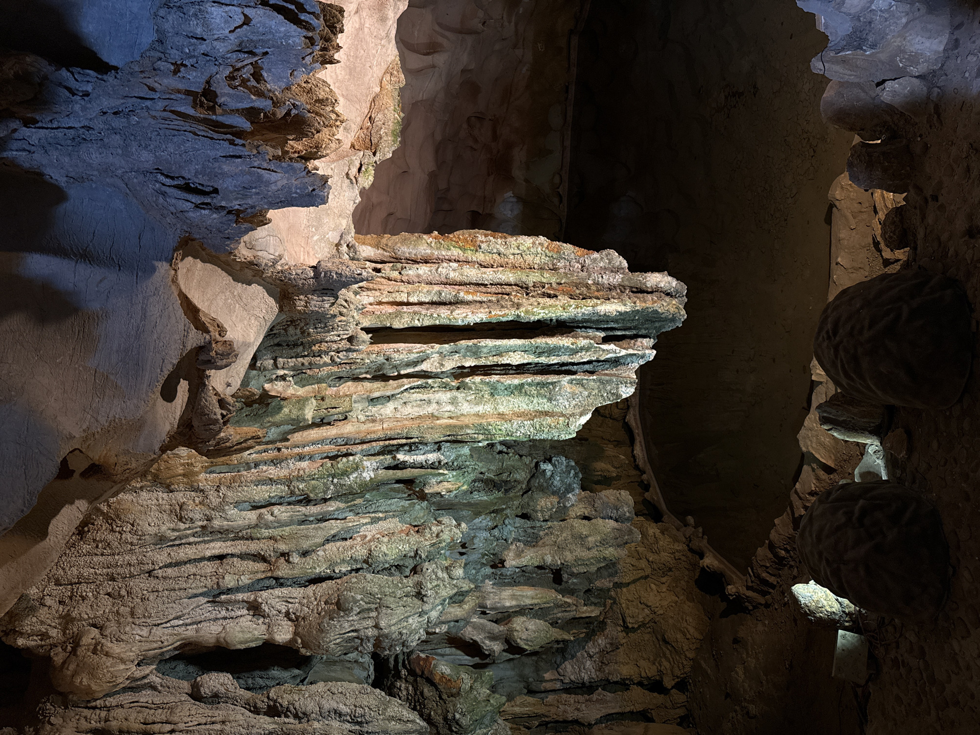 This media file presents the illuminated interior of a cave, characterized by intricate geological formations. The central subject is a prominent, horizontally layered rock structure, appearing like shelves or stratified rock, which displays various colors including greens, oranges, and whites, suggesting mineral deposits or biological growth.
The cave features diverse speleothems, including large, textured rock faces on the left, some appearing bluish-gray with complex, broken surfaces. On the right side, several large, rounded, and textured formations, reminiscent of brains or fungal growths, are visible along the cave wall. The overall texture of the cave surfaces varies significantly, from smooth and water-worn to rough, crystalline, and granular.
Artificial lighting strategically illuminates specific areas, creating dramatic contrasts of light and shadow that highlight the formations' contours and textures. Deep shadows recede into the background, hinting at the cave's vastness. No human activity or visible text is present. The scene evokes a sense of ancient, natural grandeur, typical of subterranean environments. Given the context, this cave is located in Ha Long Ward, Vietnam.