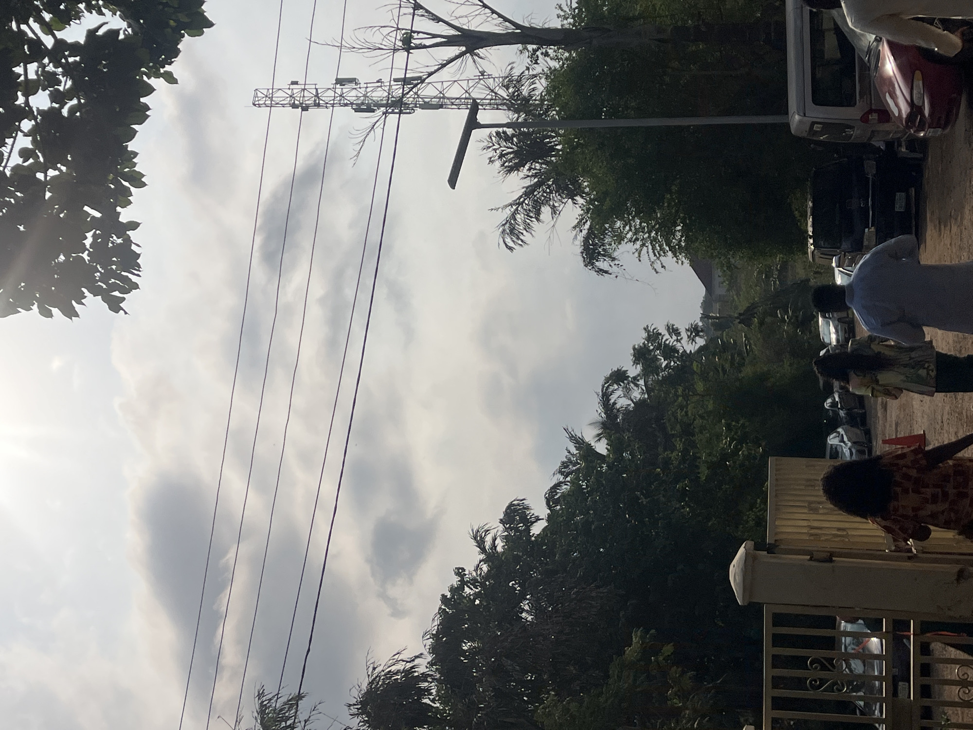 The image depicts a street scene in Galadimawa Village, Nigeria. Tall trees and a metal utility pole with multiple power lines dominate the left side of the frame, contrasting with a cloudy sky.
To the right of the frame, there is a line of parked cars, including a red car and a white van. A person wearing a light blue shirt walks near the cars. In the foreground, a portion of a metal gate is visible, with people standing behind it. The overall environment appears to be a residential area or a village street with a mix of vegetation, infrastructure, and human activity.