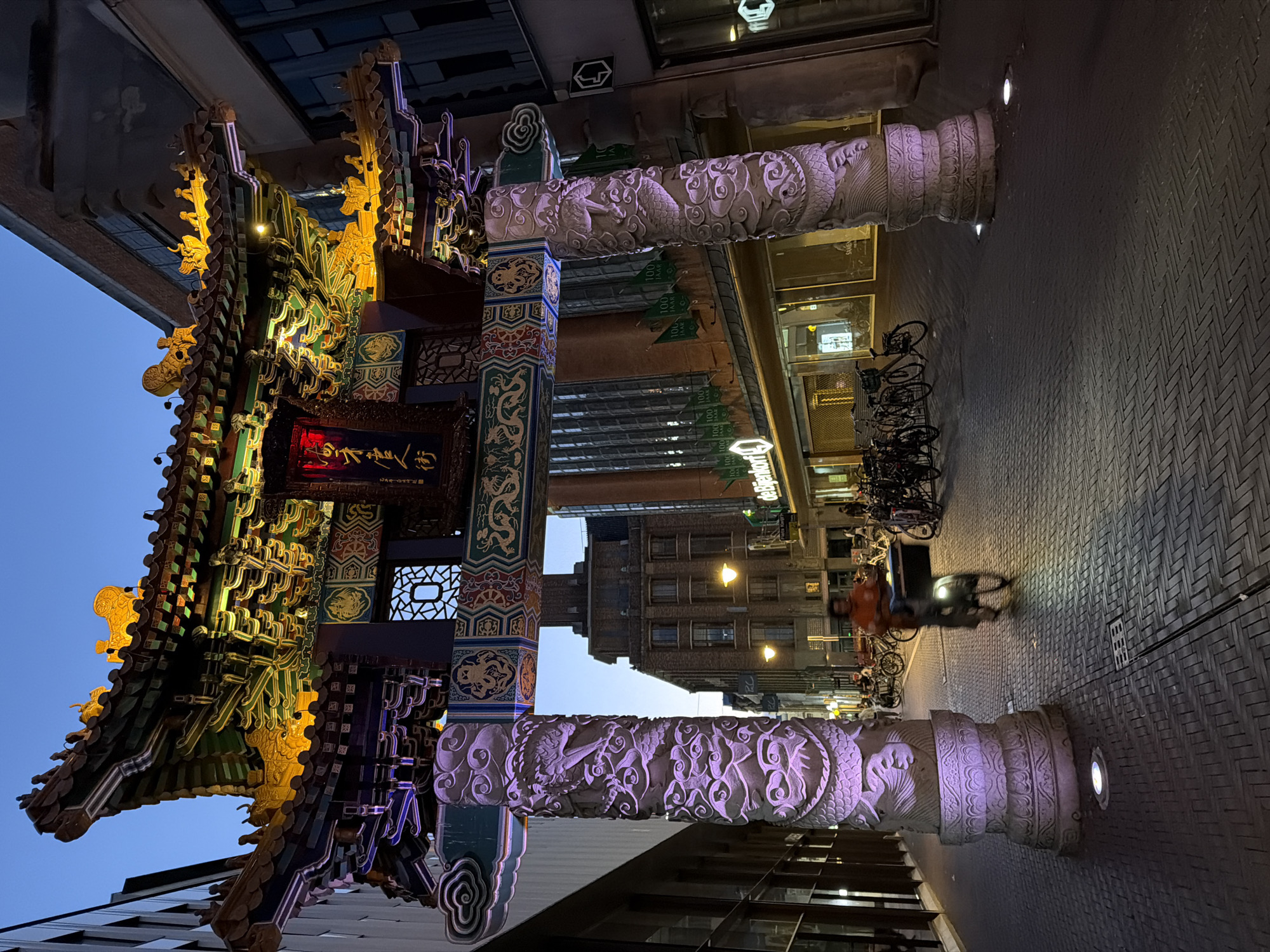 The image depicts the entrance gate to The Hague's Chinatown, located on the city's edge along the Grote Marktstraat. The gate, an ornate structure with traditional Chinese architectural elements, is illuminated by warm yellow and purple lights. The sky above is a deep twilight blue.
In the foreground, two large, intricately carved pillars support the gate. The pillar on the left is illuminated with purple light, showcasing detailed carvings of dragons and clouds. The pillar on the right is also carved with similar motifs and is primarily lit by the ambient light of the street.
A person is riding a bicycle from left to right across the paved street, their silhouette slightly blurred due to motion. The street is lined with parked bicycles on the right side of the image. The background reveals the architecture of the city, with buildings featuring large windows and storefronts. Signs for "Bijenkorf" are visible, indicating a department store. The ground is paved with brick-like tiles, arranged in a herringbone pattern. Small ground lights are also present, casting pools of light on the pavement.