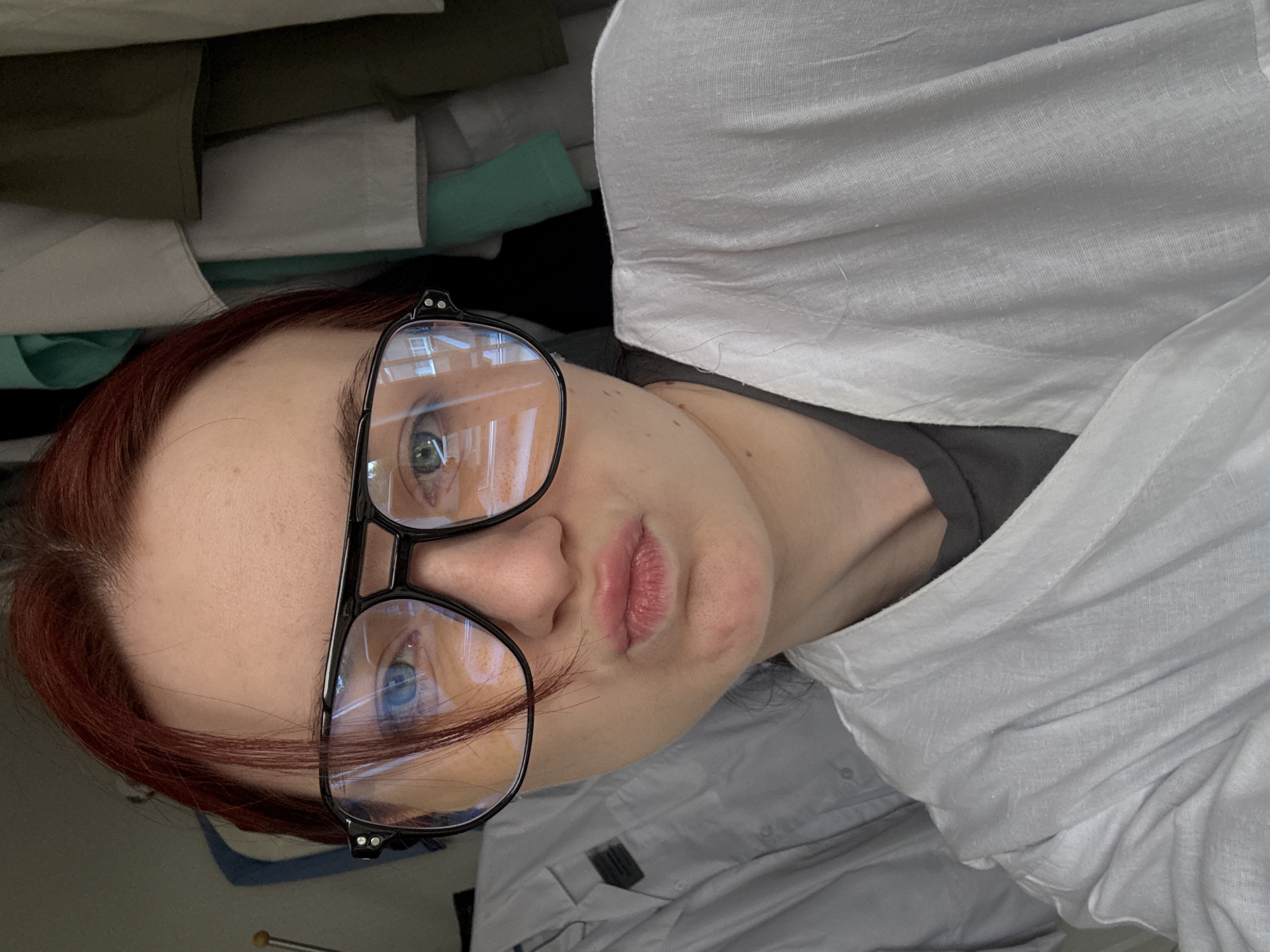 The image shows a close-up selfie of a young woman with red hair and blue eyes, wearing oversized black-rimmed glasses and a white lab coat over a dark gray shirt. She is making a pouty face. The background is cluttered with what appears to be hanging clothes, possibly in a closet or laundry room. The image does not contain any information or visual cues to suggest it was taken in Saint Petersburg, Russia.