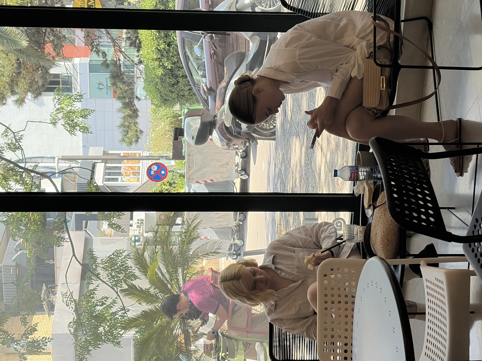 This image captures a street-level view through a window of a cafe or restaurant in Budva, Montenegro. Two women are seated at tables, one in the foreground and one further back.
In the foreground, a young woman with blonde, shoulder-length hair is seated at a black chair facing a large, round, dark table. She is wearing a light-colored button-down shirt and appears to be eating something small and yellow, possibly fruit or a pastry. Her attention is directed downwards towards her lap. A glass with a dark liquid and a straw is on the table next to her. Another chair, white with a perforated design, is positioned in front of the table.
Further back, another young woman with her hair in a bun is seated at a black chair at a smaller table. She is wearing a white button-down shirt and is looking down at her smartphone, which she holds in her hands. A light-brown handbag with a strap is placed on the chair beside her. A bottle of water and a small ceramic cup are on the table in front of her.
The background visible through the window reveals a street scene. A man in a pink shirt is seated at a table in the mid-ground, looking at his phone. Palm trees and other greenery are prominent. Beyond the immediate street area, buildings of varying heights and architectural styles are visible, including a modern white building with a logo that appears to be "MLADI BUDVE" along with colorful accents and windows. Traffic signals and metal refuse bins on wheels are also present. A dark-colored scooter or motorcycle is parked in front of one of the buildings. The overall impression is a sunny day, with strong natural light illuminating the scene.