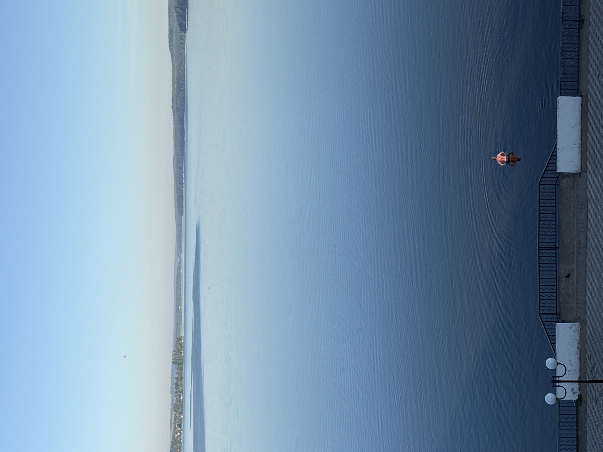 The image depicts a body of water, likely a lake or river, in Votkinsk, Russia. The water is calm, with gentle ripples and a gradient of color from light to dark blue. A person is swimming in the water, relatively close to a concrete embankment. The embankment features a black metal railing and white concrete blocks. Streetlights are visible along the embankment. In the distance, the opposite shore is visible, lined with trees and some buildings. The sky is clear and light blue.