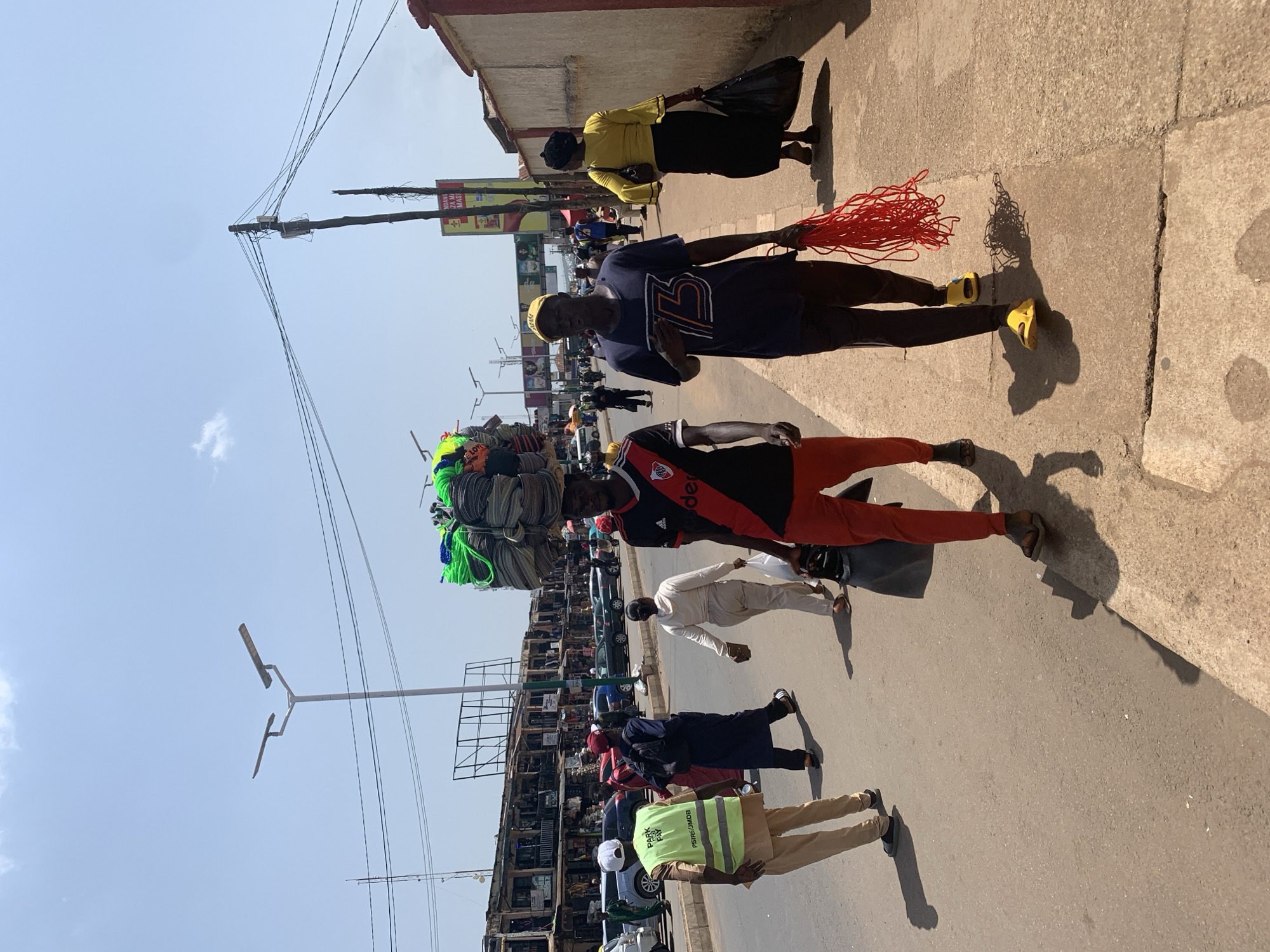 This is a street scene in Jos, Nigeria, on a bright, sunny day. A man is walking on the sidewalk, carrying a bundle of bright green and blue ropes on his head. He is wearing a red and black soccer jersey and red pants. To his left, another man walks beside him, wearing a dark blue t-shirt with a white logo and black pants. He is holding a large bundle of red rope in his right hand. In the background, other pedestrians and vehicles are visible on the street, and buildings line the opposite side of the road, suggesting a market area. The overall impression is of a bustling urban environment.