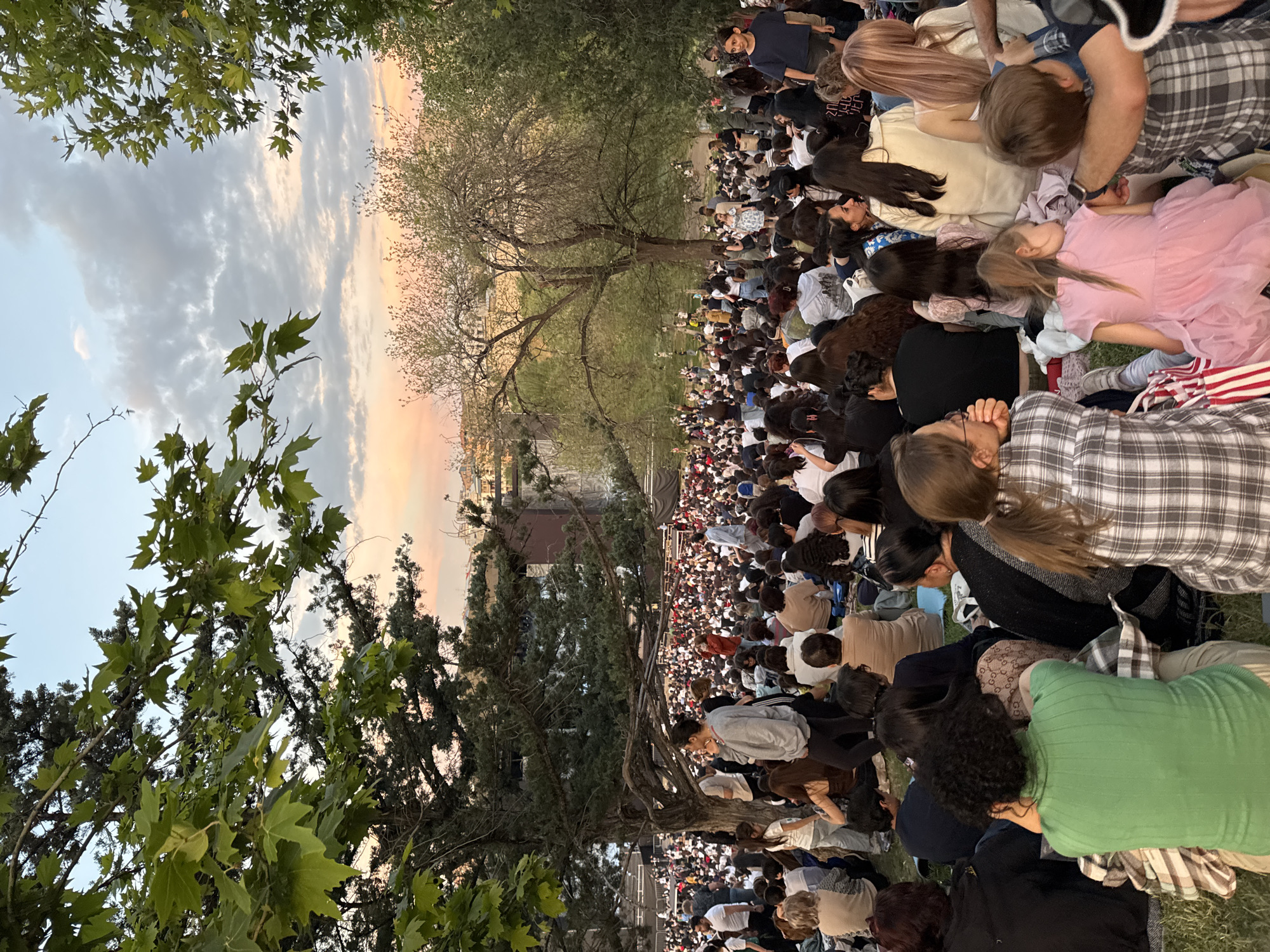This image captures a large outdoor gathering on a grassy slope in the Cascade Complex in Yerevan, Armenia, during twilight. Hundreds, possibly thousands, of individuals are seated or standing on the descending green terrain, oriented towards a distant, unseen focal point.
In the immediate foreground, several people are visible from behind, including a person in a green ribbed long-sleeve top seated on a plaid blanket, a young girl in a pink dress, and a young boy in a plaid shirt. Various blankets and personal items are spread on the grass.
The midground is dominated by a dense crowd extending down the slope. The individuals are casually dressed, with a notable prevalence of dark hair among the observed attendees. Large trees with green foliage are interspersed within the crowd and frame the scene on both sides, including a prominent tree with a thick trunk and extensive branches on the left, and another large, partially defoliated tree further back on the right.
The background reveals a large, multi-tiered structure, likely part of the Cascade Complex or an associated performance stage, with minimal visible detailing due to the distance and lighting. Beyond this structure, the cityscape of Yerevan unfolds under a sky transitioning from pale blue to soft orange and pink, indicative of sunset, with scattered grey clouds. The overall lighting is diffuse and warm.