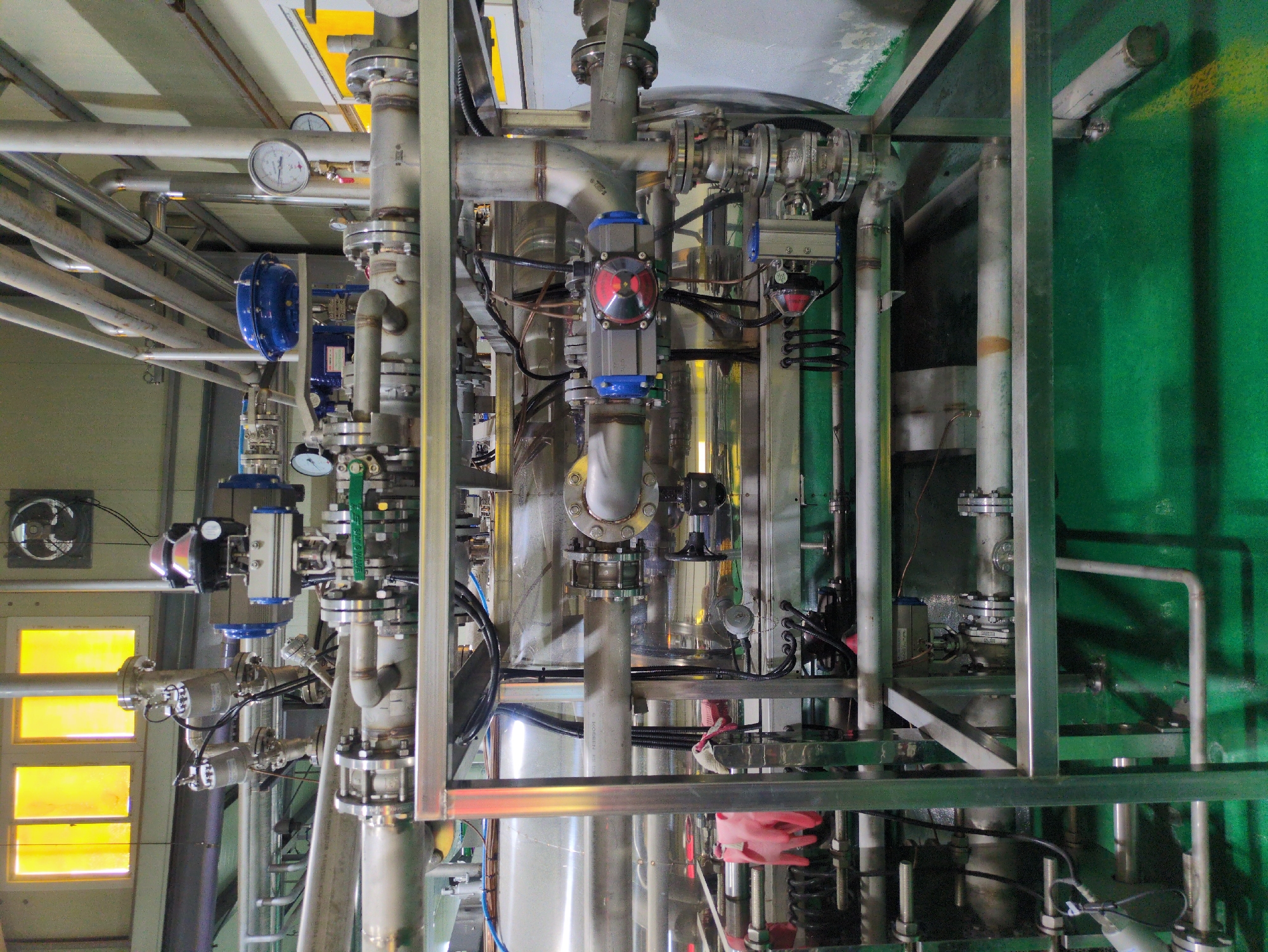 The image depicts an industrial setting, likely inside a chemical processing plant or similar facility, located in an unknown city. The scene is dominated by complex stainless steel piping and equipment, including valves, pressure gauges, and what appears to be a large cylindrical tank. Some of the valves are fitted with automated actuators. The pipes are supported by a network of metal framing. The background includes a wall painted green and a window with yellow panes. The overall impression is one of a highly engineered, functional space designed for industrial processes. There is no evidence of human presence in the immediate scene.