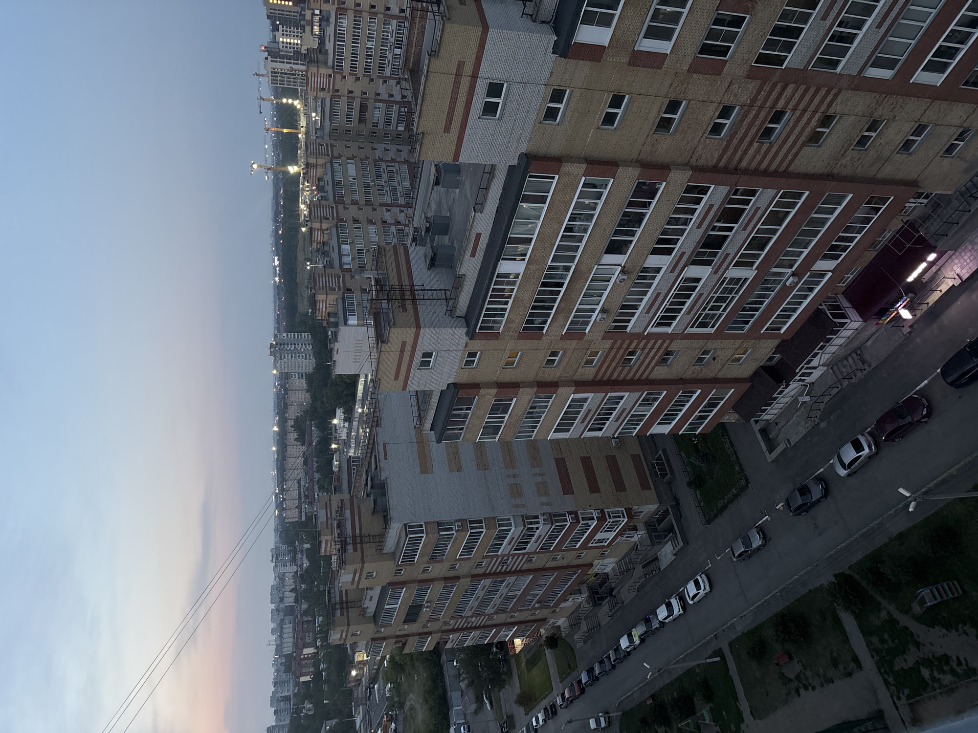 This is an aerial, medium-angle, daylight shot of a residential area in Omsk, Russia. The scene depicts multi-story apartment buildings, some of which are under construction with cranes visible in the distance. The sky is a pale blue with a hint of pink from the setting sun. In the foreground, a street lined with parked cars runs alongside one of the apartment buildings. The architecture is typical of Soviet-era mass housing, characterized by concrete structures and repetitive window patterns. The overall atmosphere is one of urban development and everyday life in a Russian city.