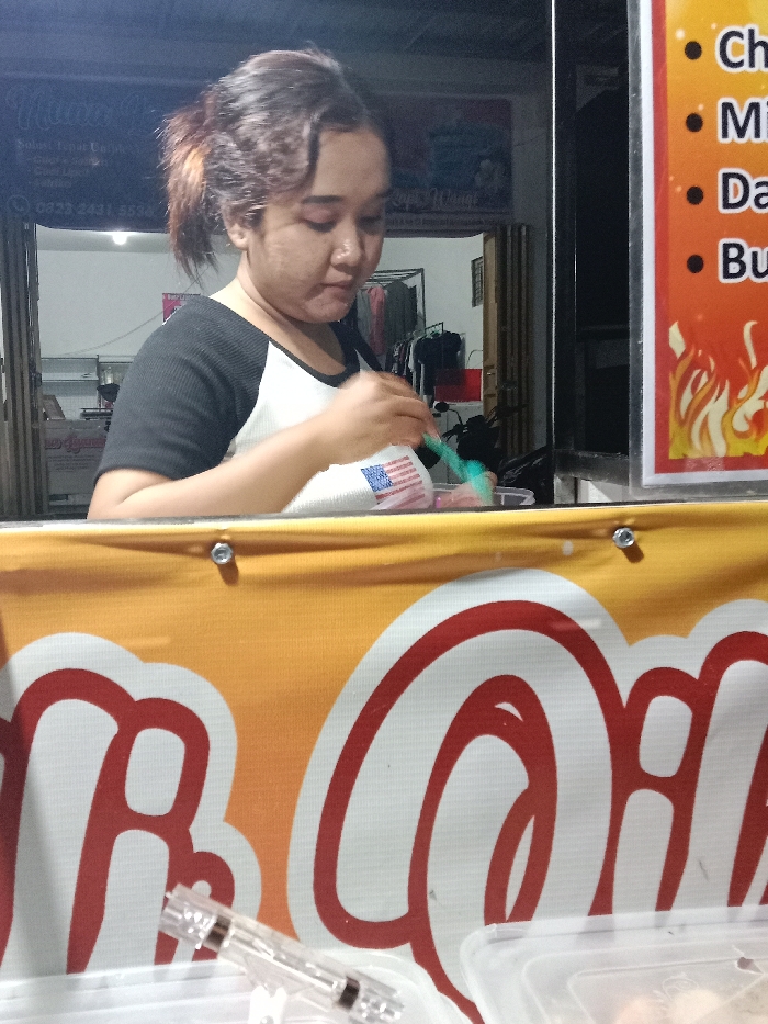 The image depicts a female individual engaged in food preparation within what appears to be a street food stall or small eatery, located in Turunsih, Indonesia. The female, with brown hair tied back, wears a black and white raglan-style short-sleeved shirt. She is observed stirring or mixing contents in a clear plastic container using a teal spoon. The container features a design resembling the flag of the United States.

In the immediate foreground, a yellow and orange banner with partial white and red lettering is visible, secured by two silver fasteners. On a surface beneath this banner, a clear plastic syringe-like object and another clear plastic food container are present.

The background behind the female reveals an adjacent storefront or entrance. To the left, a dark-colored facade displays white text, including the partially visible word "Menu" and the phone number "0823 2431 5536". Inside this area, clothing hangs on a rack, and a dark motorcycle is parked. To the female's right, a vertical menu board with an orange background and red flame graphics at the bottom lists menu items such as "Ch," "Mi," "Da," and "Bu" under black bullet points. The scene is illuminated by artificial light, suggesting an evening or nighttime setting.