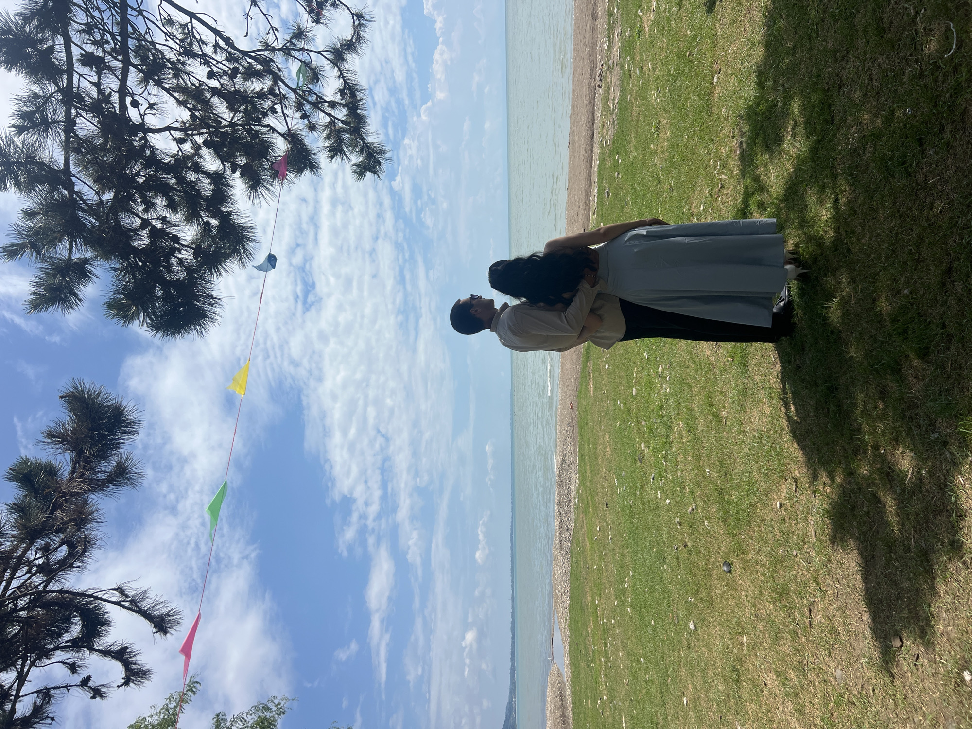 In an outdoor scene, a man and a woman are standing on a grassy area facing a body of water under a bright, cloudy sky. The man, wearing a tan collared shirt and dark pants, has his right arm around the woman's shoulders. The woman, with long dark hair, is wearing a light blue, floor-length skirt and a matching top. A string of colorful triangular flags is visible in the sky between pine tree branches. The shoreline is composed of sand and pebbles, meeting a large expanse of water.
