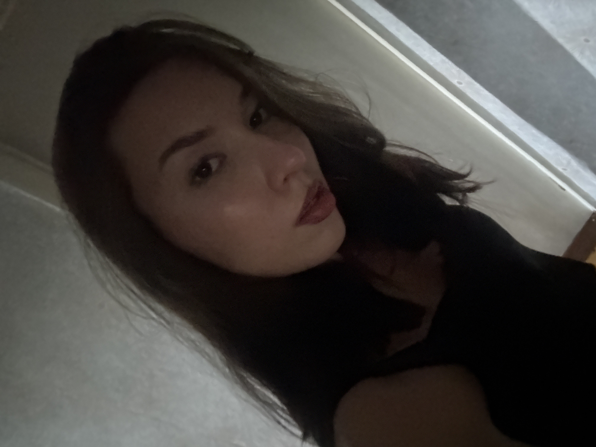 A young woman with dark hair and red lipstick is captured in a dimly lit indoor setting. She is looking towards the camera with a neutral expression, her face partially illuminated by an unseen light source. Her hair is dark and appears to be styled down, framing her face. She is wearing a black garment, likely a top or dress, that is mostly obscured by shadow. The background is indistinct, appearing to be a wall with vertical lines that could suggest a door or a window frame. There are no other individuals or objects of note in the frame. The overall atmosphere of the image is somber and intimate due to the low lighting and close-up framing. The location is not specified in the provided image or hints, therefore any details about the city of Inskoy, Russia cannot be included.