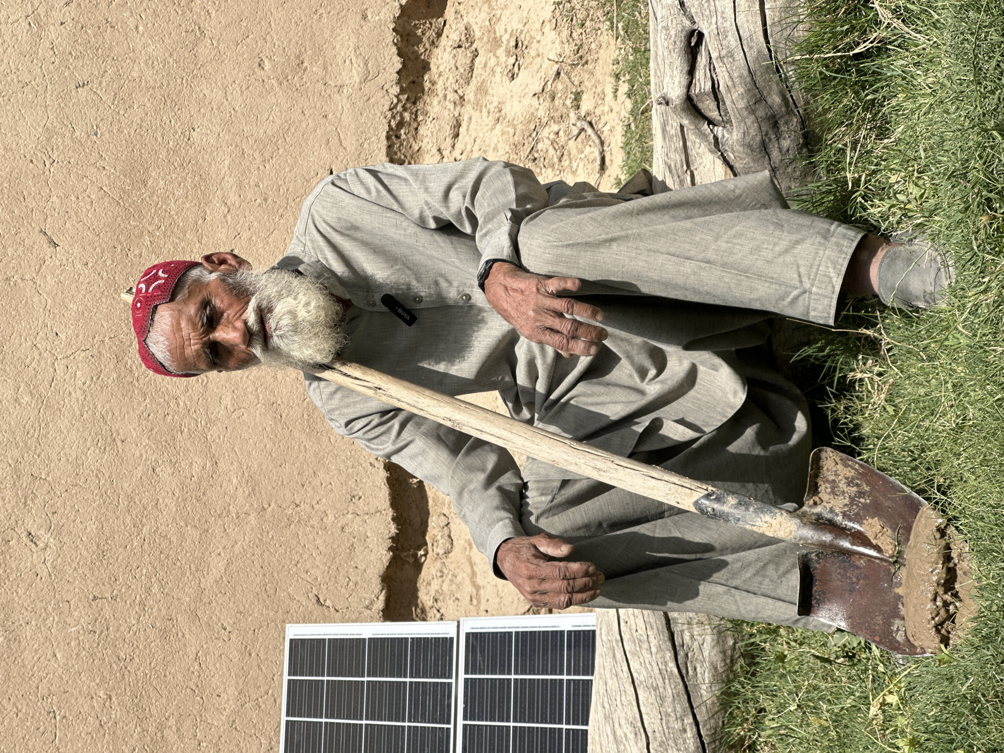 In Quetta City Tehsil, Pakistan, an elderly man sits on the ground beside a tan wall and a solar panel. He wears a red and white patterned hat, light gray clothing, and dark shoes. He has a long, white beard. He is holding a shovel covered in dirt. He is sitting next to some grass and a log.