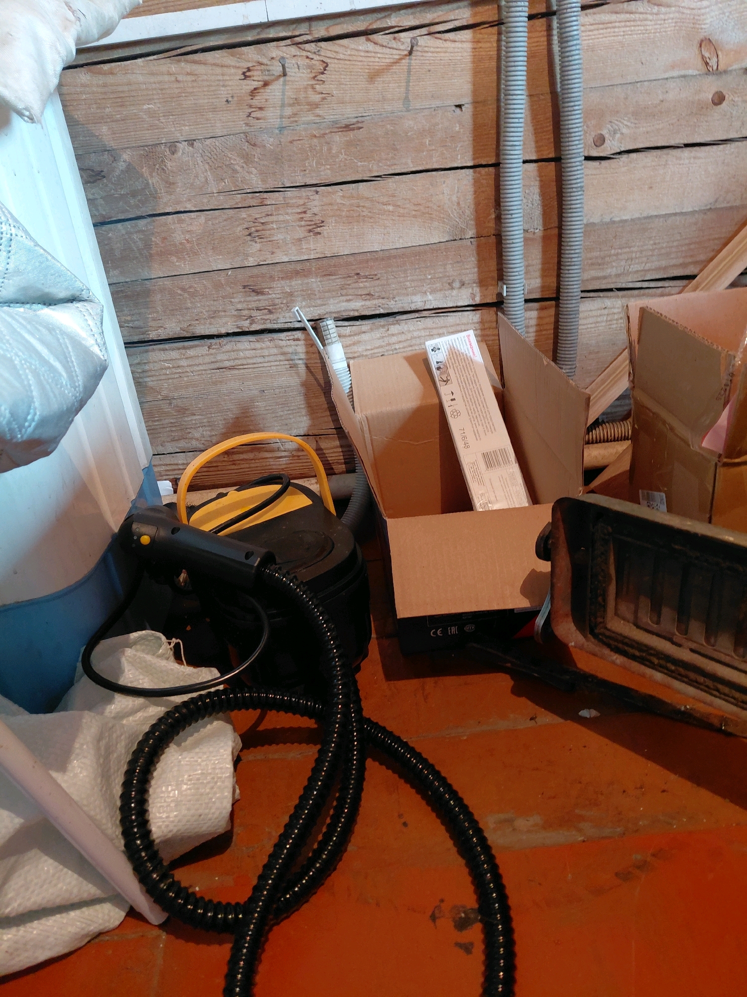 The image depicts a cluttered storage area with wooden plank walls. On the floor, various items are present including a black steam cleaner with a yellow handle and hose, a white textured bag, several cardboard boxes, and a metal object resembling a stove part. Two grey corrugated pipes are mounted on the wall. The scene appears to be indoors, possibly in a basement or utility room. No people are visible. The location is stated as Malinovka, Russia.