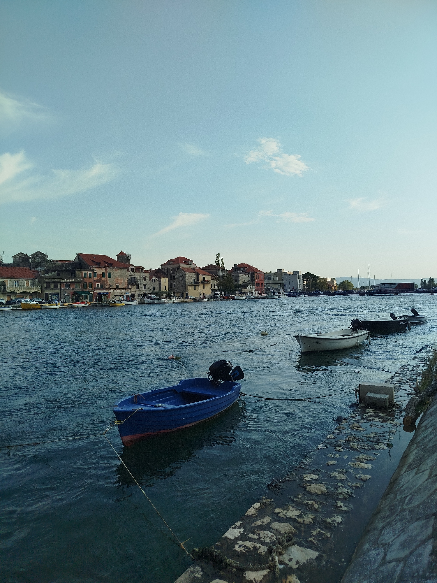 In Omiš, Croatia, a calm, blue body of water, likely a river or a sheltered bay, reflects the soft light of a partly cloudy sky. Along the far bank, a row of buildings with terracotta roofs lines the waterfront. These buildings appear to be a mix of residential and commercial establishments, with shops visible at street level. Several small boats are moored in the water. In the foreground, a bright blue boat with a black outboard motor is tied to a mooring line. Further out, two other boats, one white and one dark, are also visible. The water's edge on the right side of the image is a stone embankment. The scene suggests a tranquil day in a coastal town.
